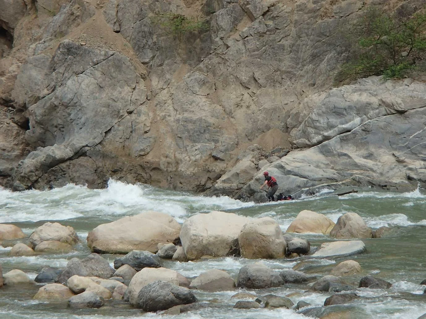 A person in a red life jacket and helmet is navigating a rocky river in a kayak, surrounded by large rocks and steep canyon walls.