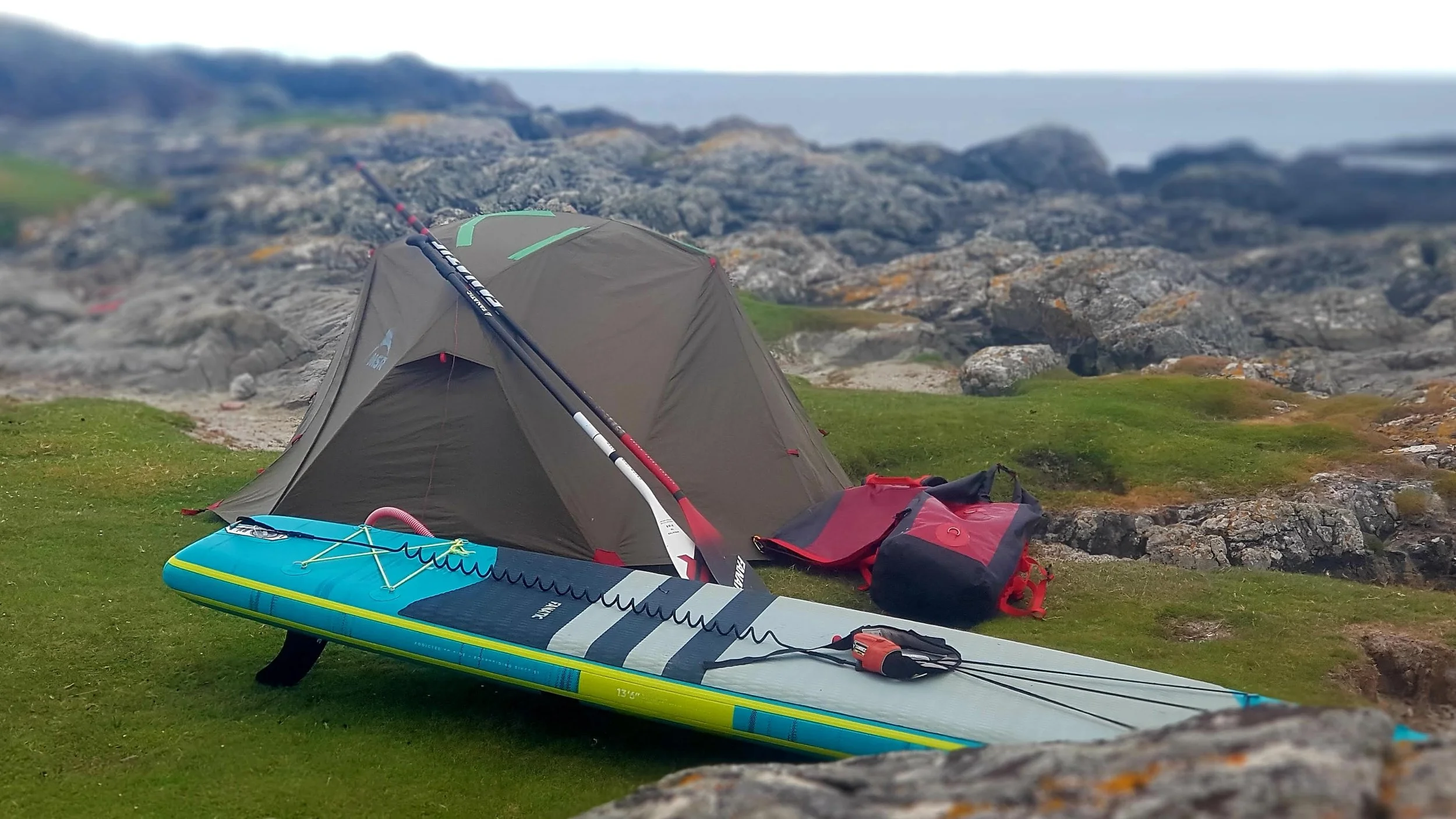 A camping tent set up near rocky terrain on grass, with paddleboard, paddles, and gear nearby, overlooking a rocky coastline in the background.