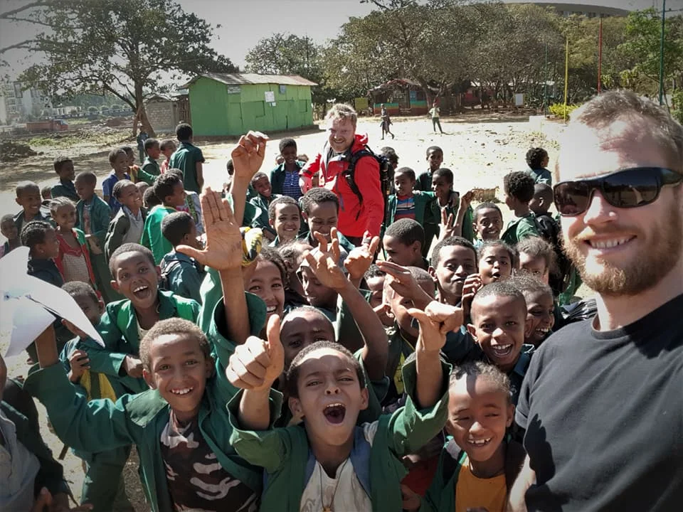 Two men taking a selfie with a large group of smiling children outdoors, with trees and a green building in the background.