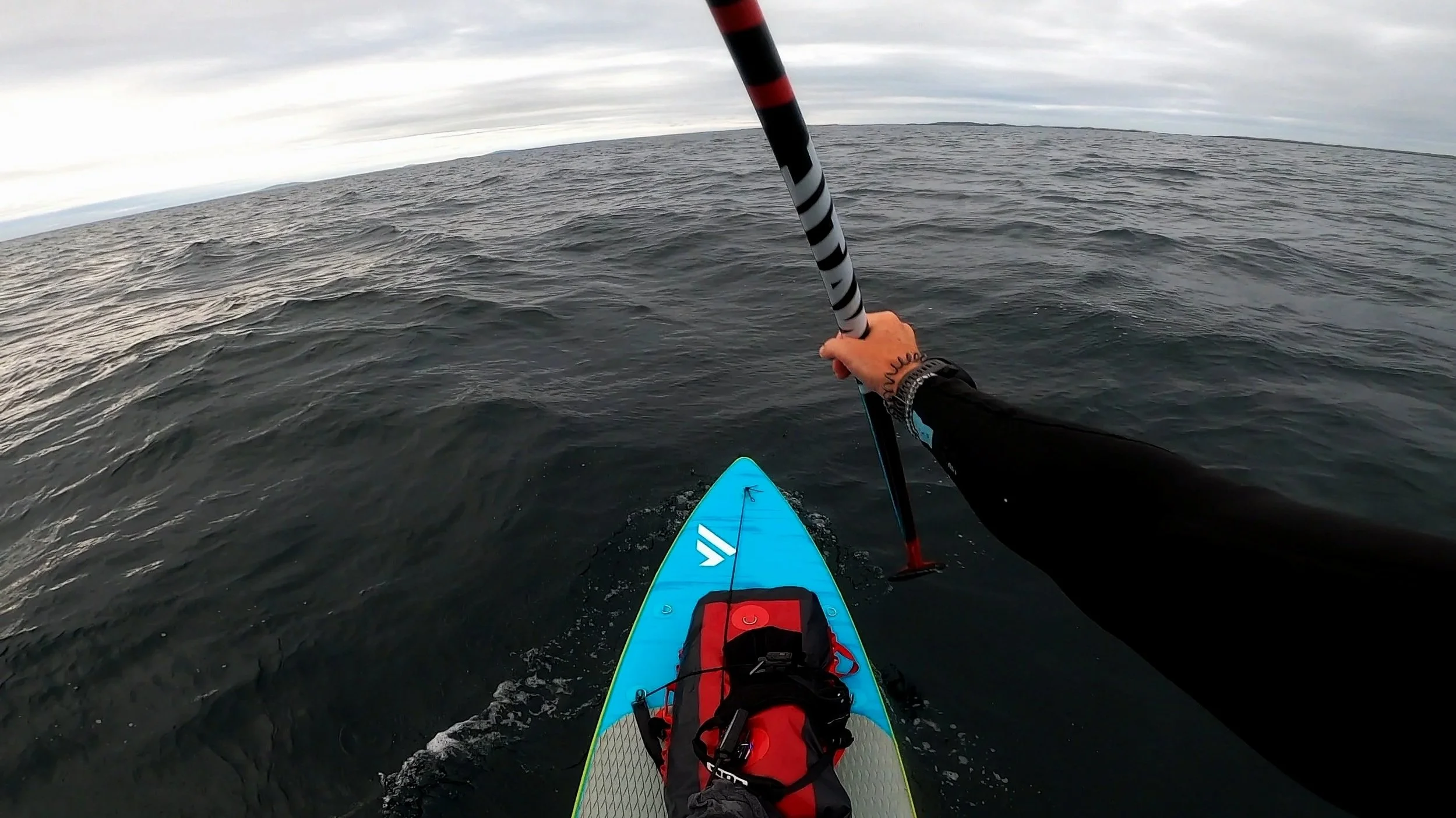 A person stand-up paddleboarding on open water with a striped paddle, wearing a black wetsuit, and holding the paddle, with a cloudy sky overhead and distant land on the horizon.