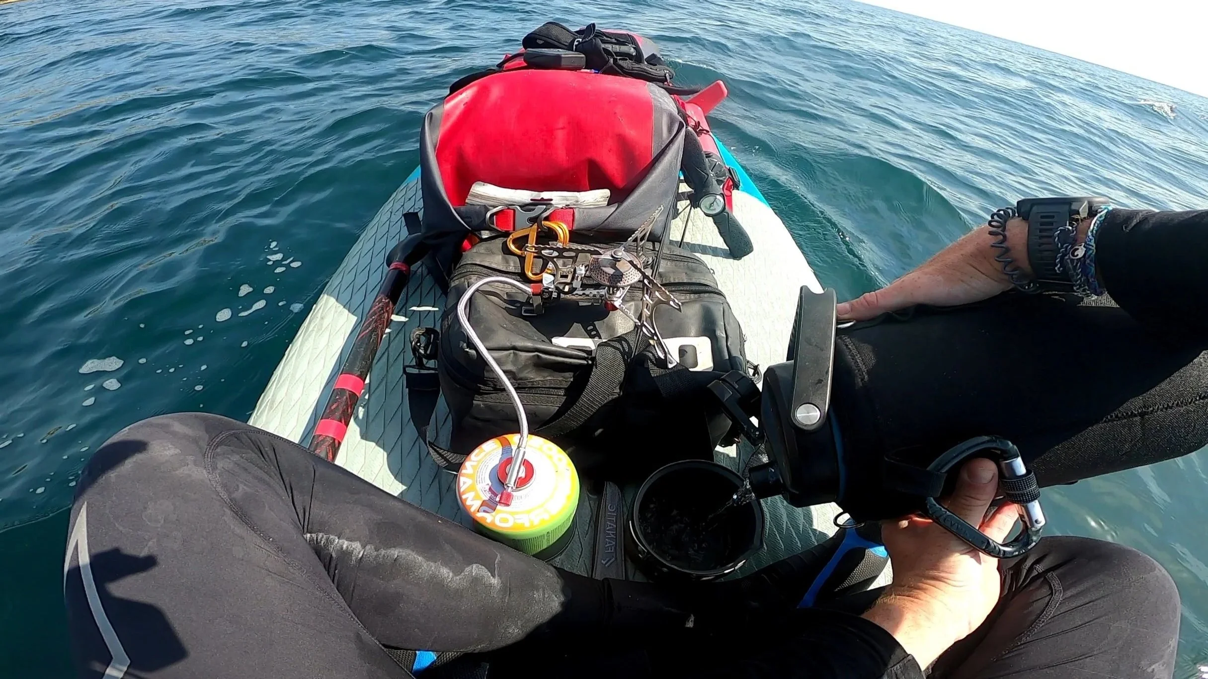 View from a paddleboard showing a person's legs and hands, with gear secured on the board, surrounded by ocean water.