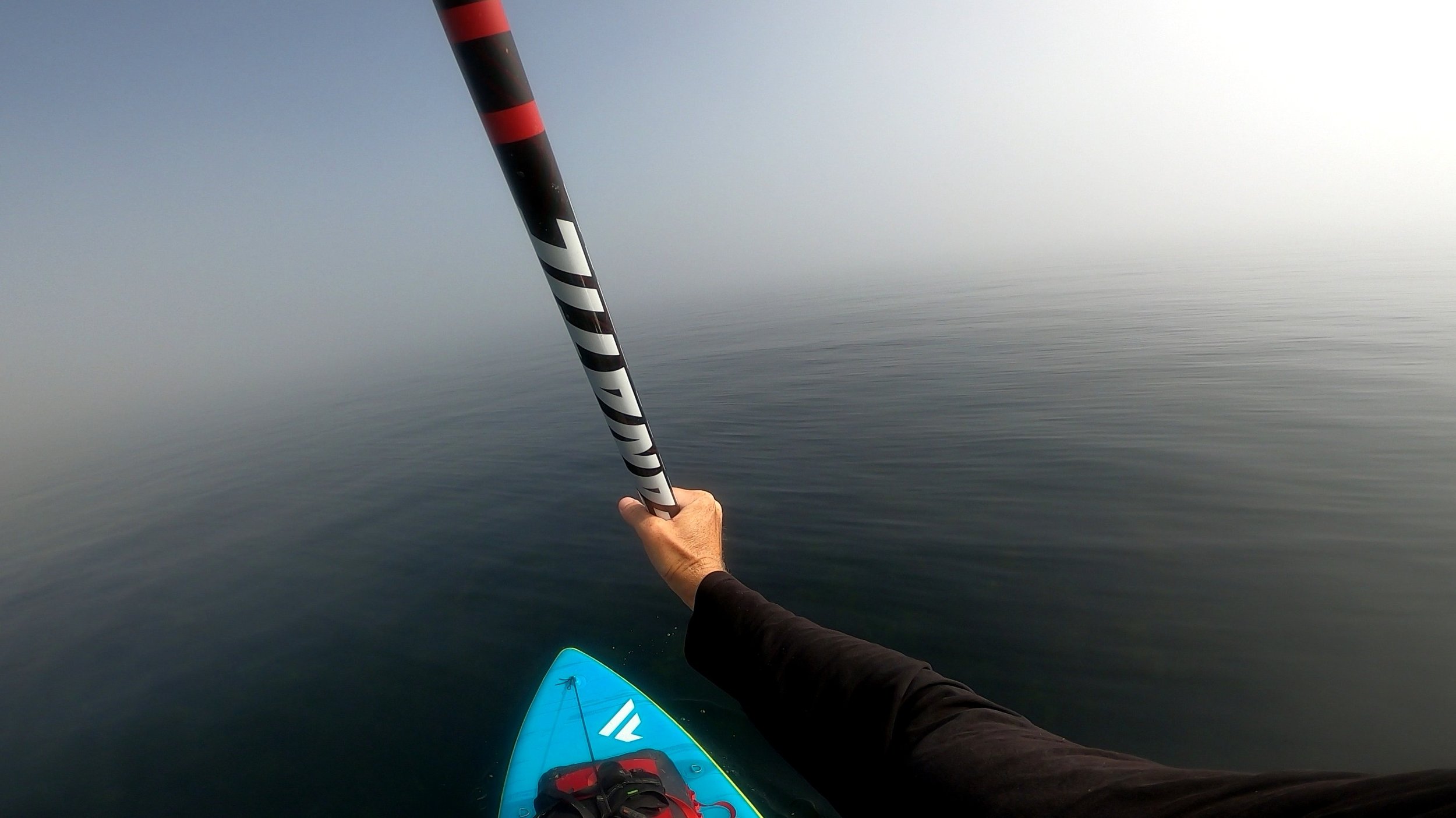 Person holding a paddle on a stand-up paddleboard on calm water, with a foggy or overcast sky.