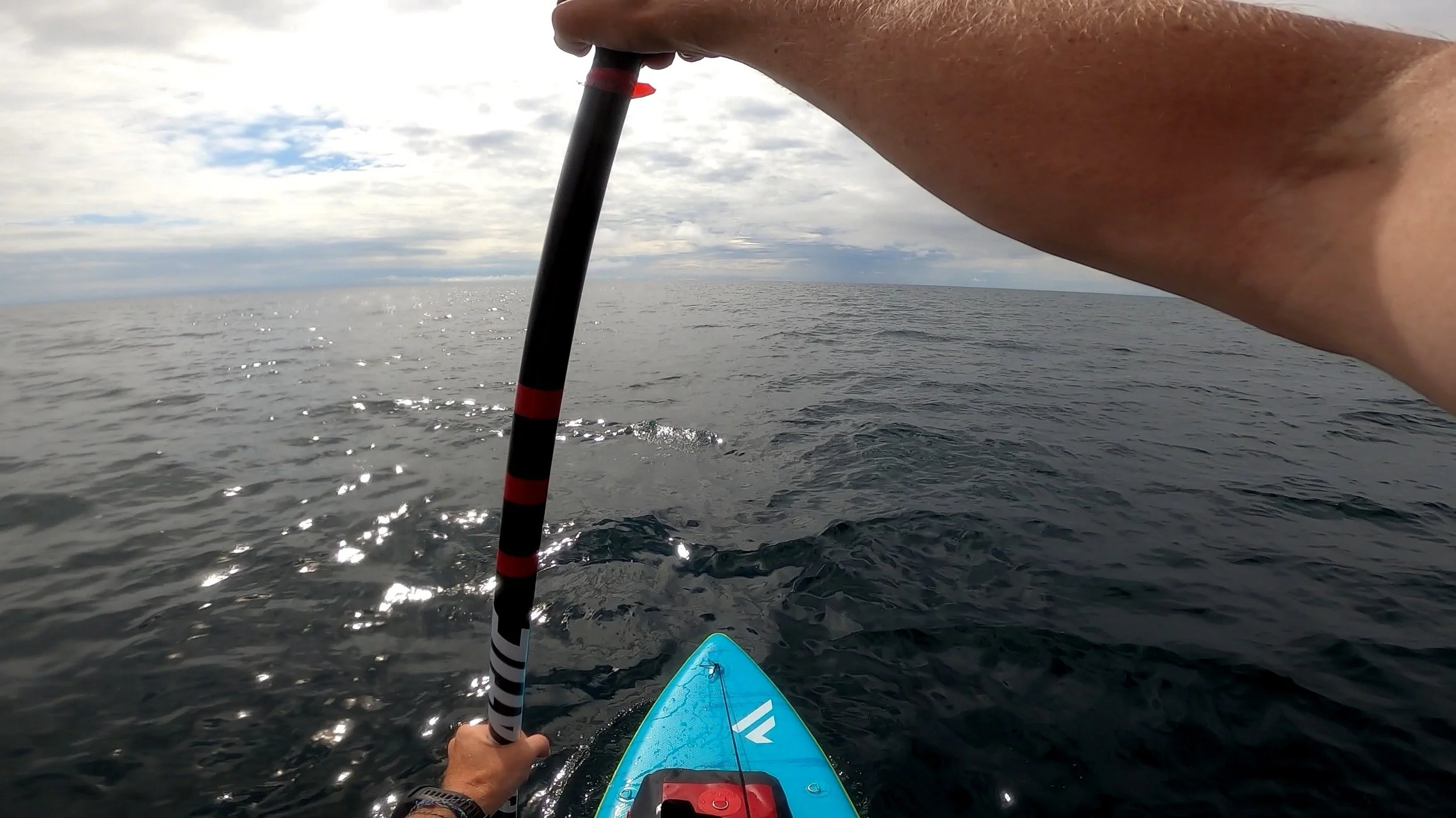 A person paddleboarding on open water during a cloudy day, with one arm holding a paddle and a portion of the paddleboard visible.