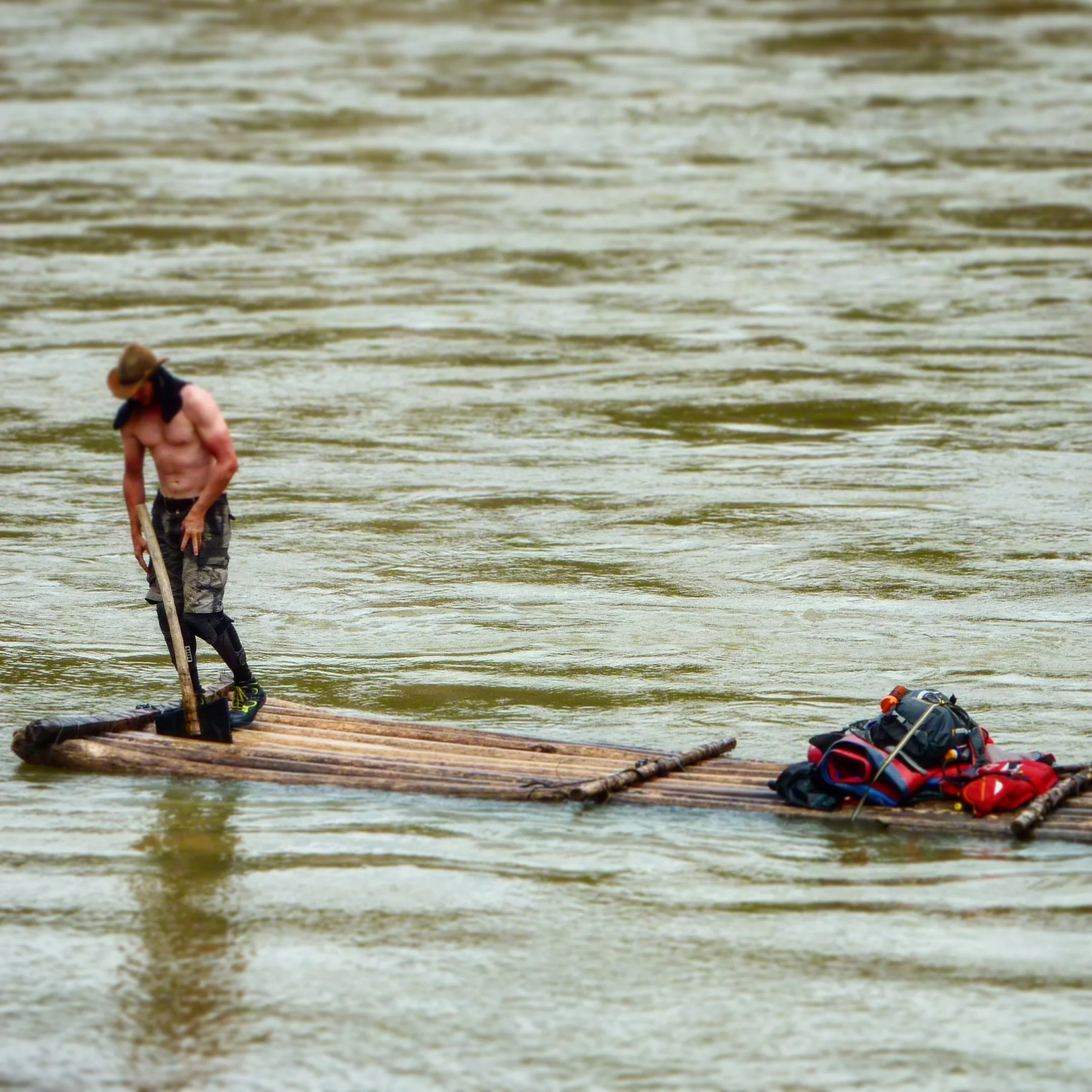 Charlie in a hat and shorts paddling a small wooden raft in a body of water. There are bags and other items on the raft.