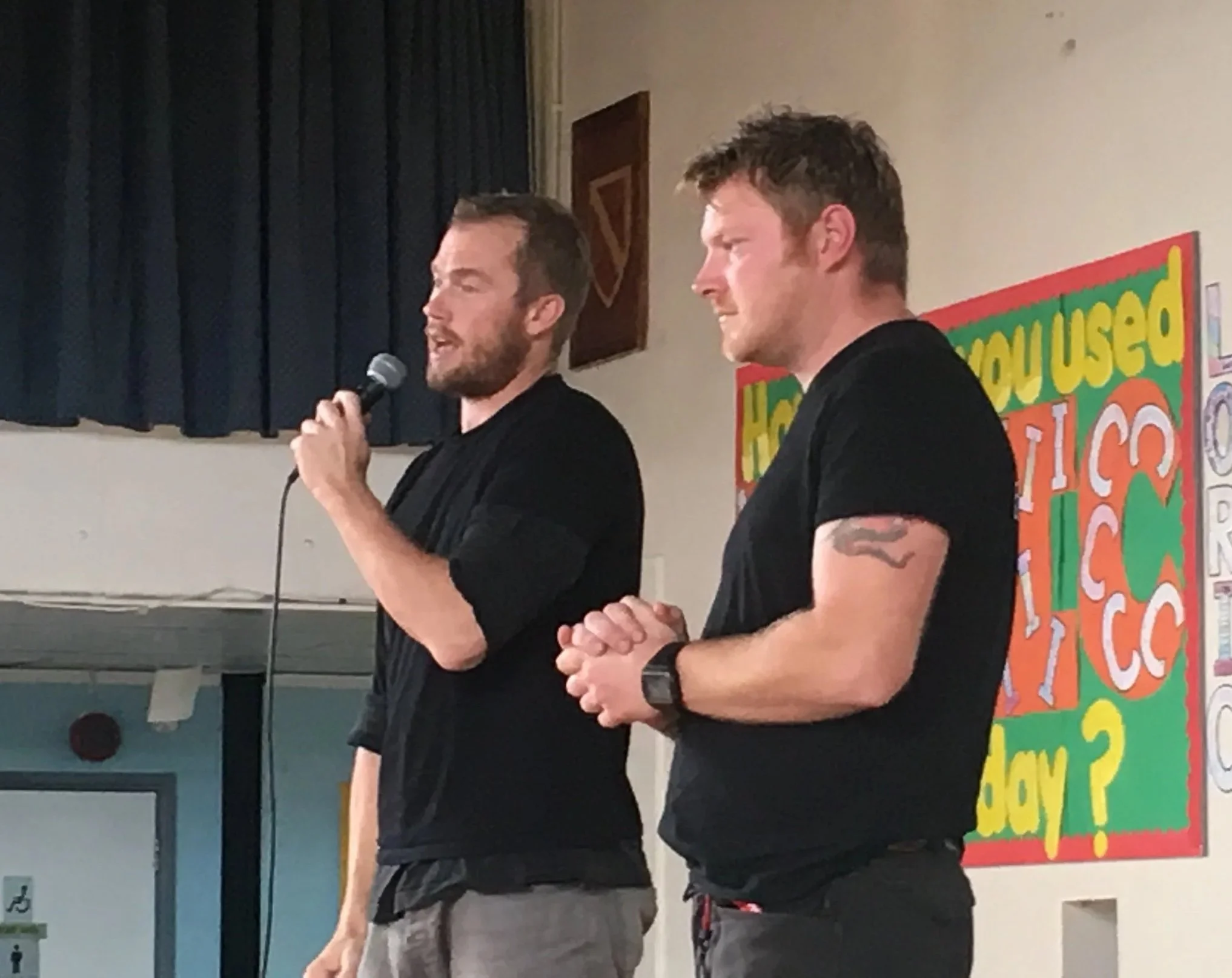 Two men standing and speaking at an indoor event, one holding a microphone, with a colorful school-themed bulletin board in the background.