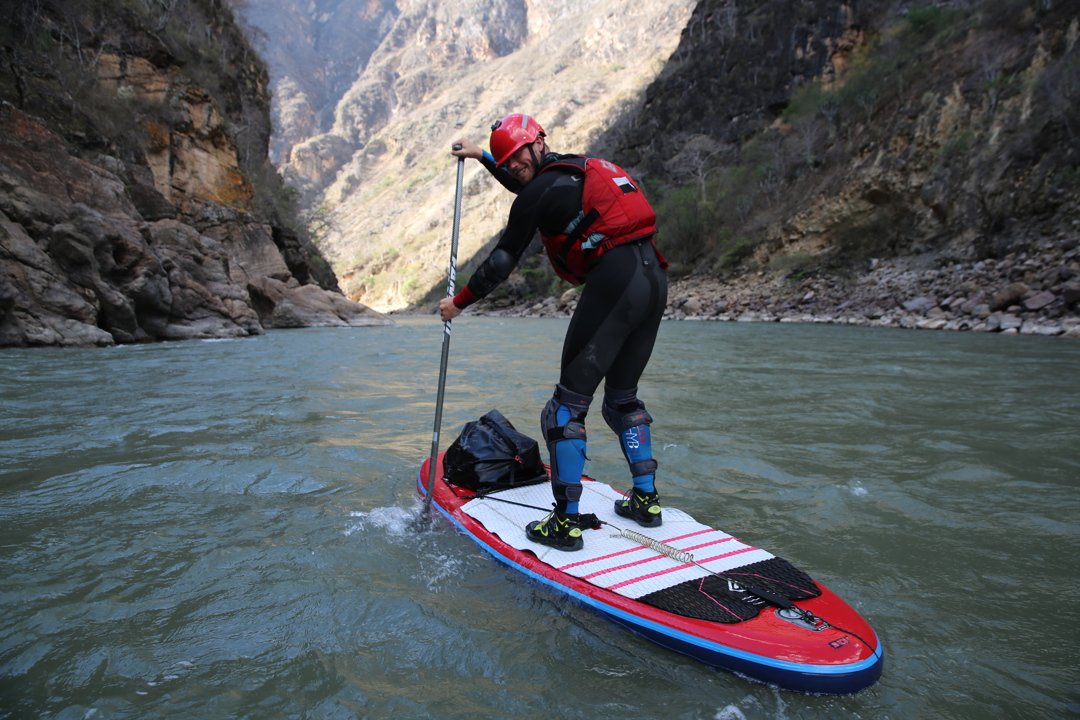 A person paddleboarding on a river in a canyon, wearing a red helmet and life vest, with mountains in the background.