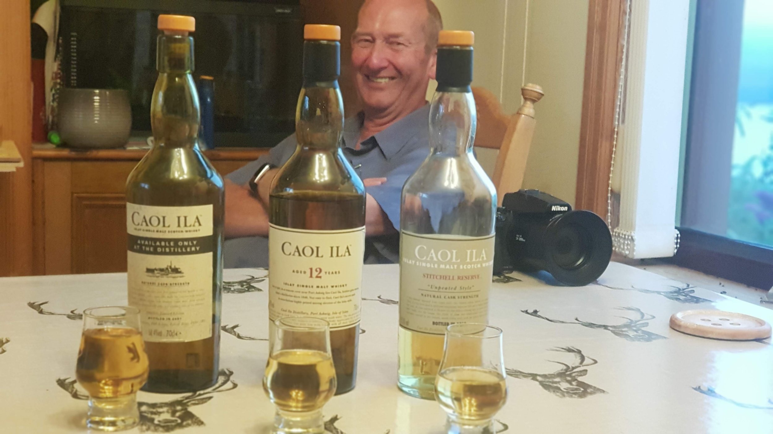 A man sitting at a dining table with three bottles of Caol Ila Scotch whisky, a camera, and glasses of whisky. The man is smiling and appears to be enjoying the moment, with a window and a cabinet in the background.