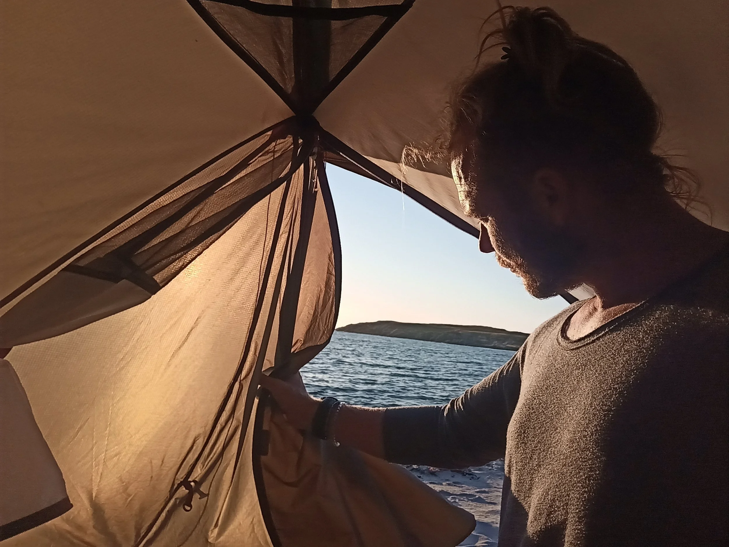 Person sitting inside a tent near a body of water at sunset.