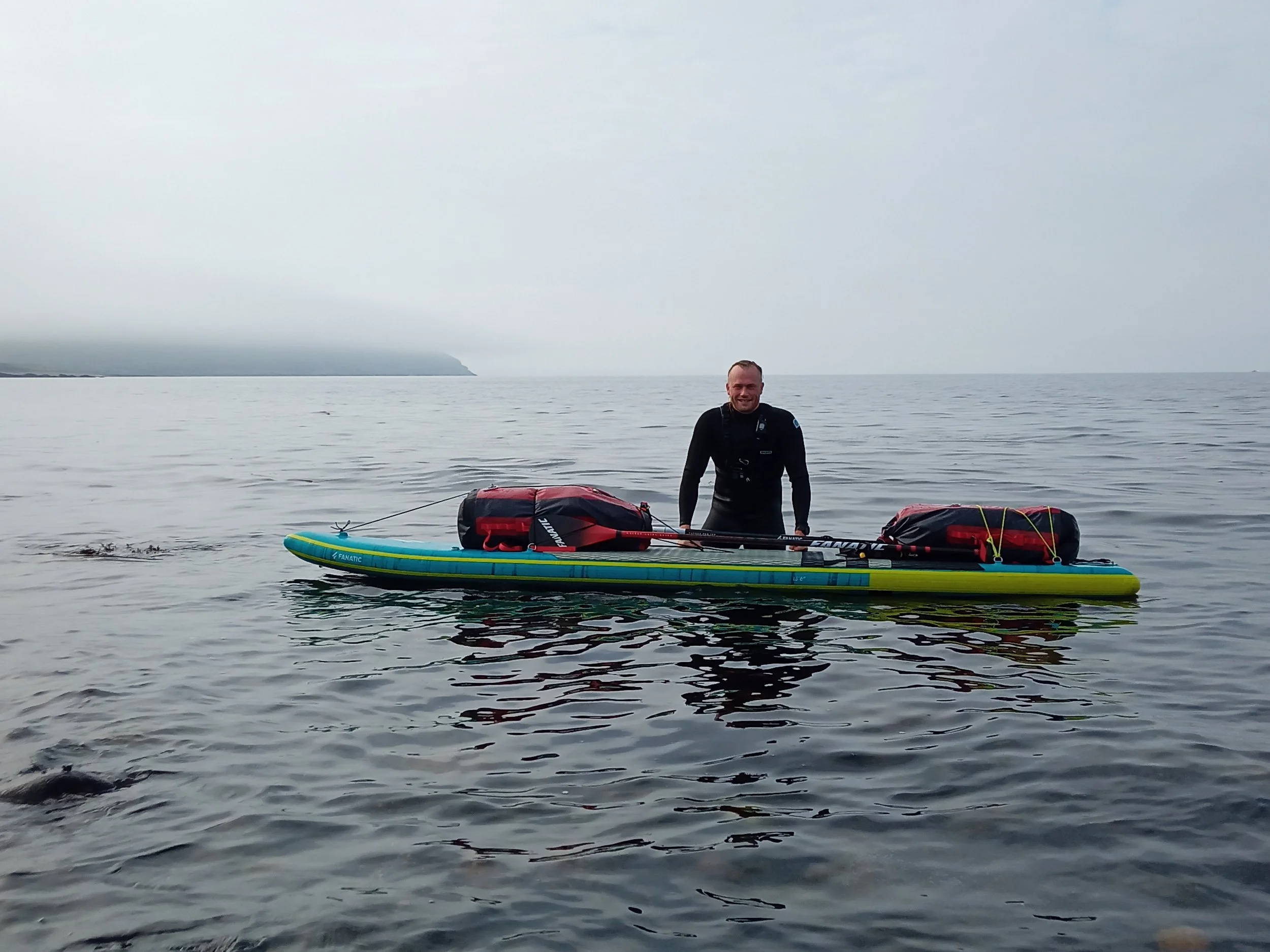 A man standing on a paddleboard in the water, smiling, with overcast skies and a distant shoreline in the background.