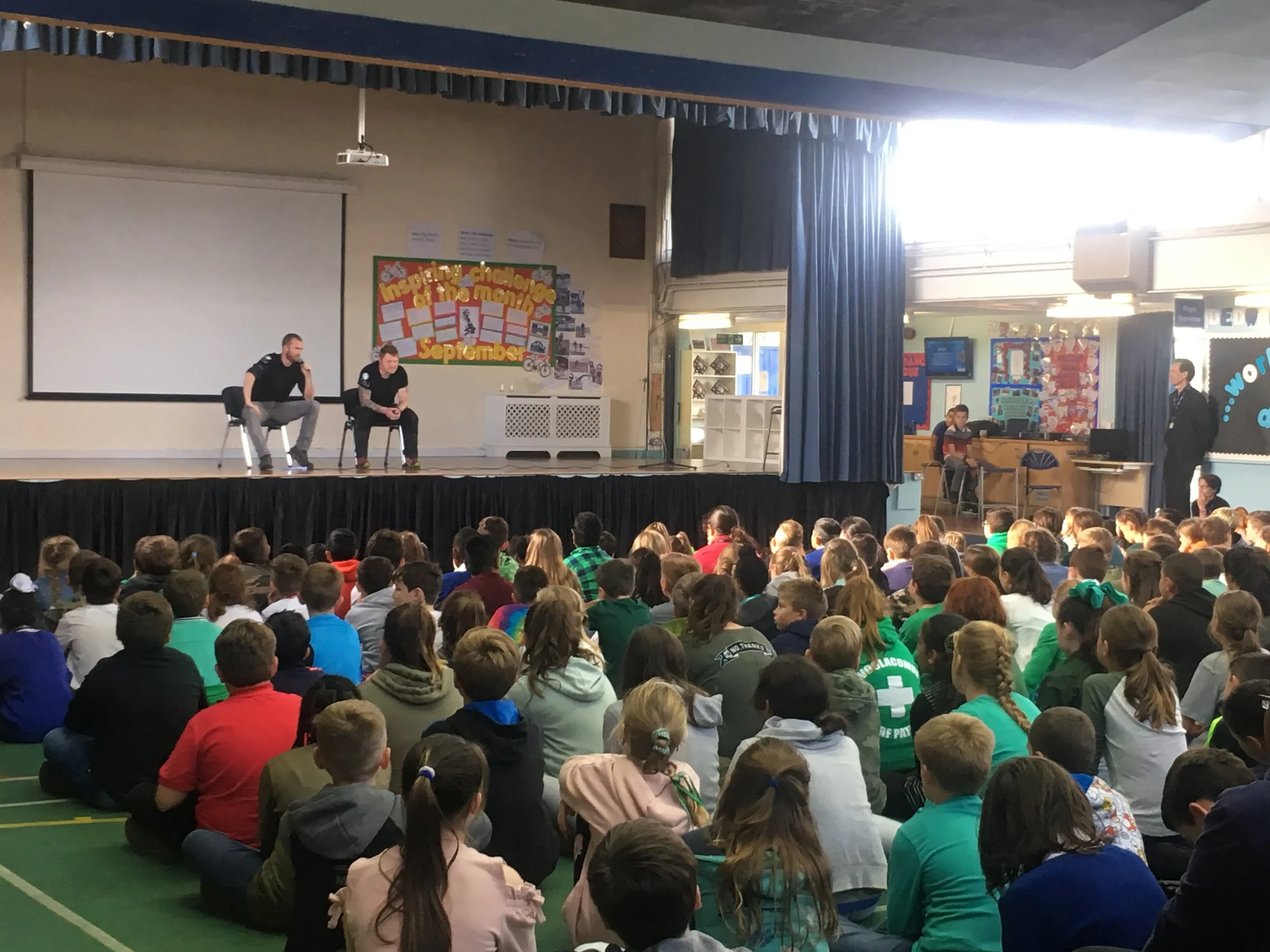 A school assembly with students seated on the floor watching a stage where two men are seated, one with a microphone speaking. The backdrop displays a colorful poster for a September themed event, and there are teachers and students seated around in a gymnasium decorated with banners and posters.
