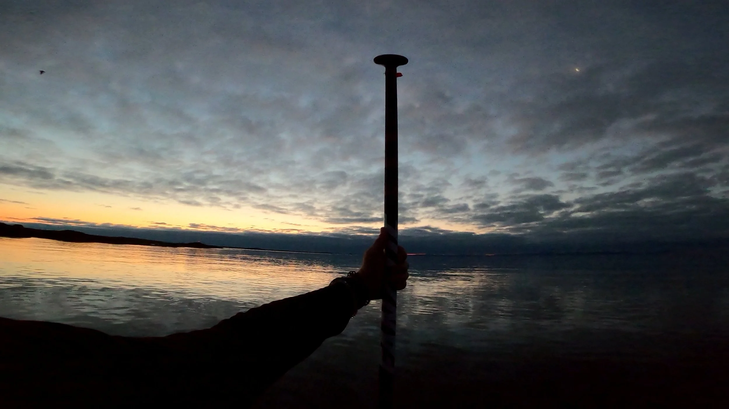 Person holding a striped staff or pole near a body of water during sunset or sunrise, with clouds and a faint star or planet visible in the sky.