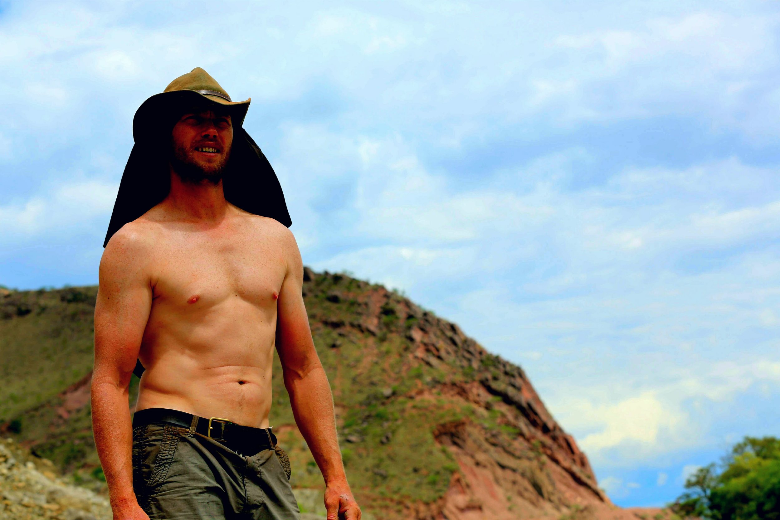 A shirtless Charlie wearing a cowboy hat and camouflage shorts standing outdoors against a backdrop of a hill and a cloudy sky.