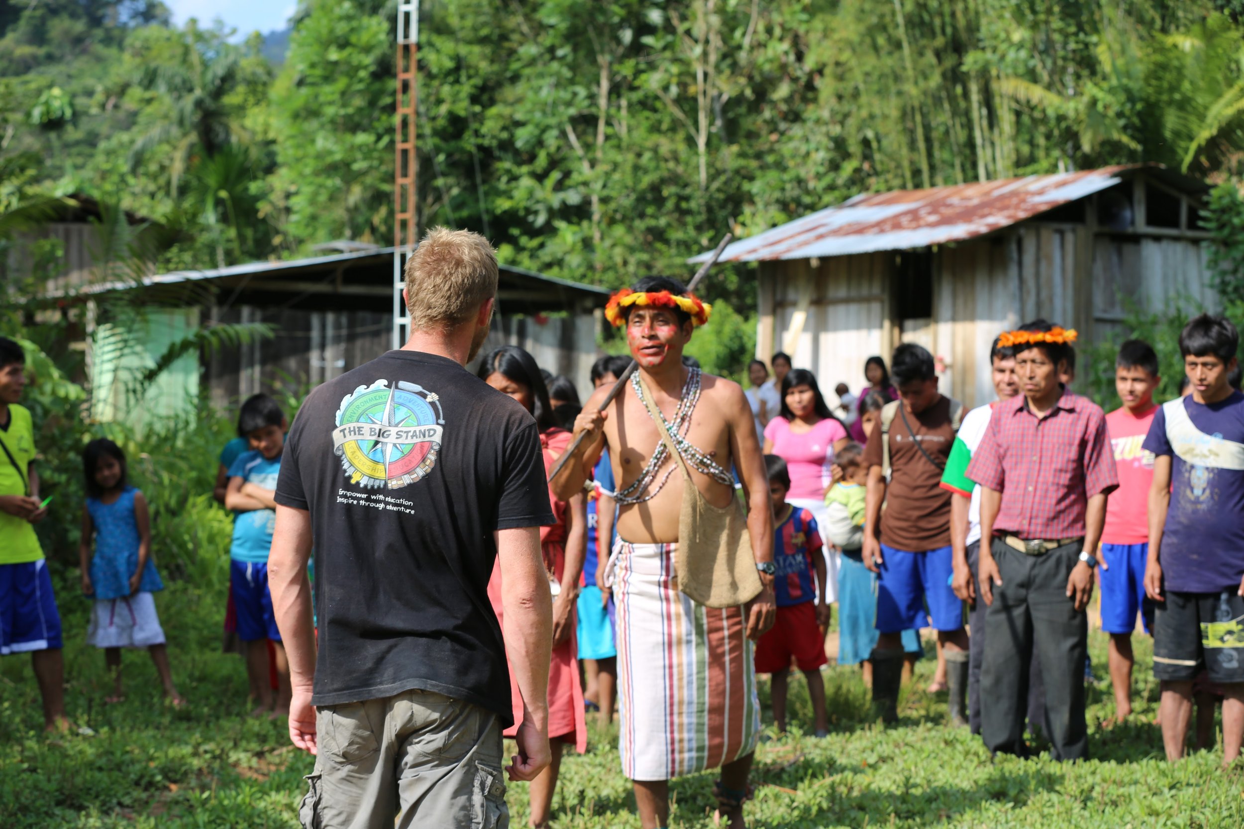A man with face paint and traditional attire, holding a staff, conversing with a person in a black t-shirt in a rural area with a group of children and adults, surrounded by lush green trees and wooden buildings.