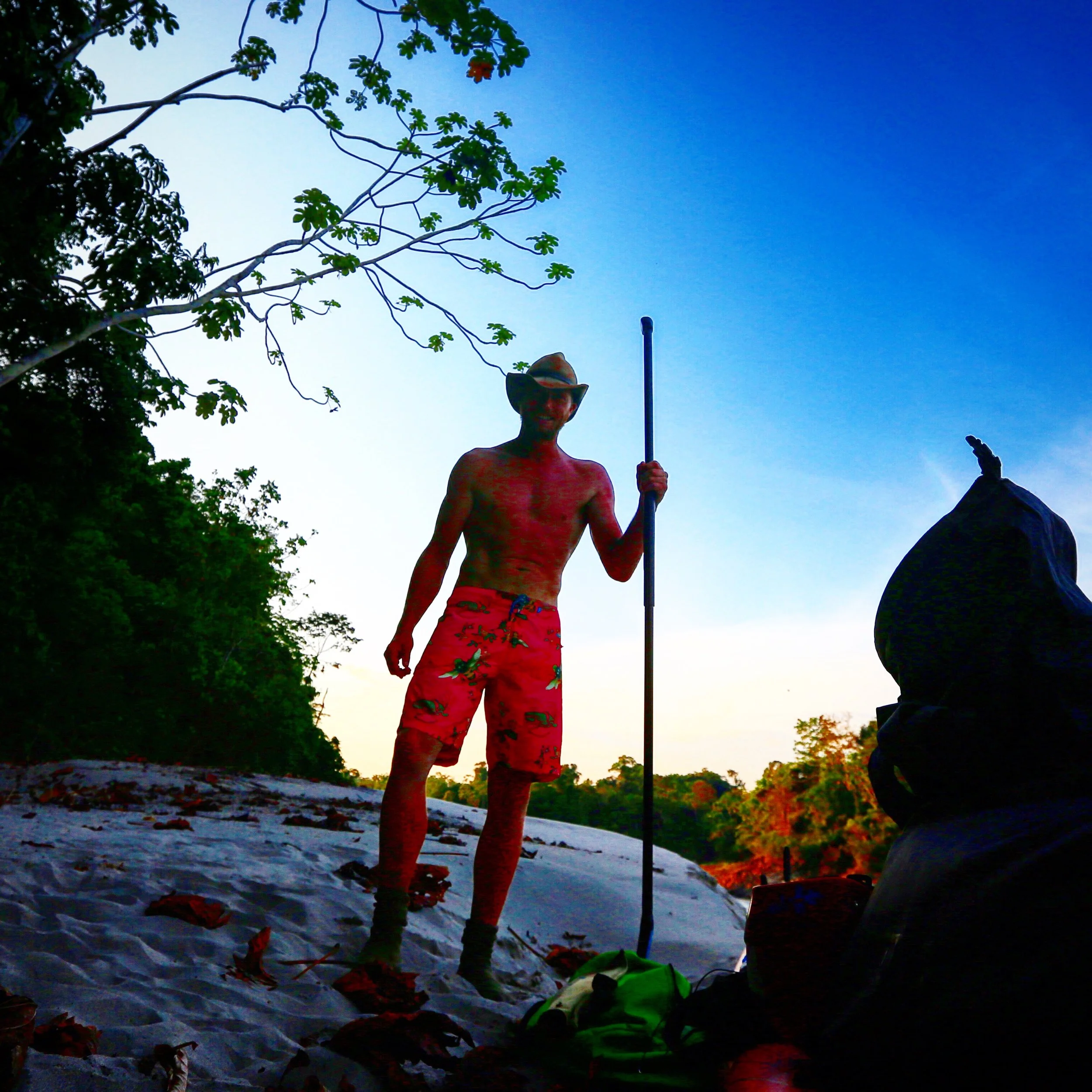 A man wearing a hat and red floral swim trunks standing in a rural outdoor setting during sunset, holding a long stick, with trees and a clear blue sky in the background.