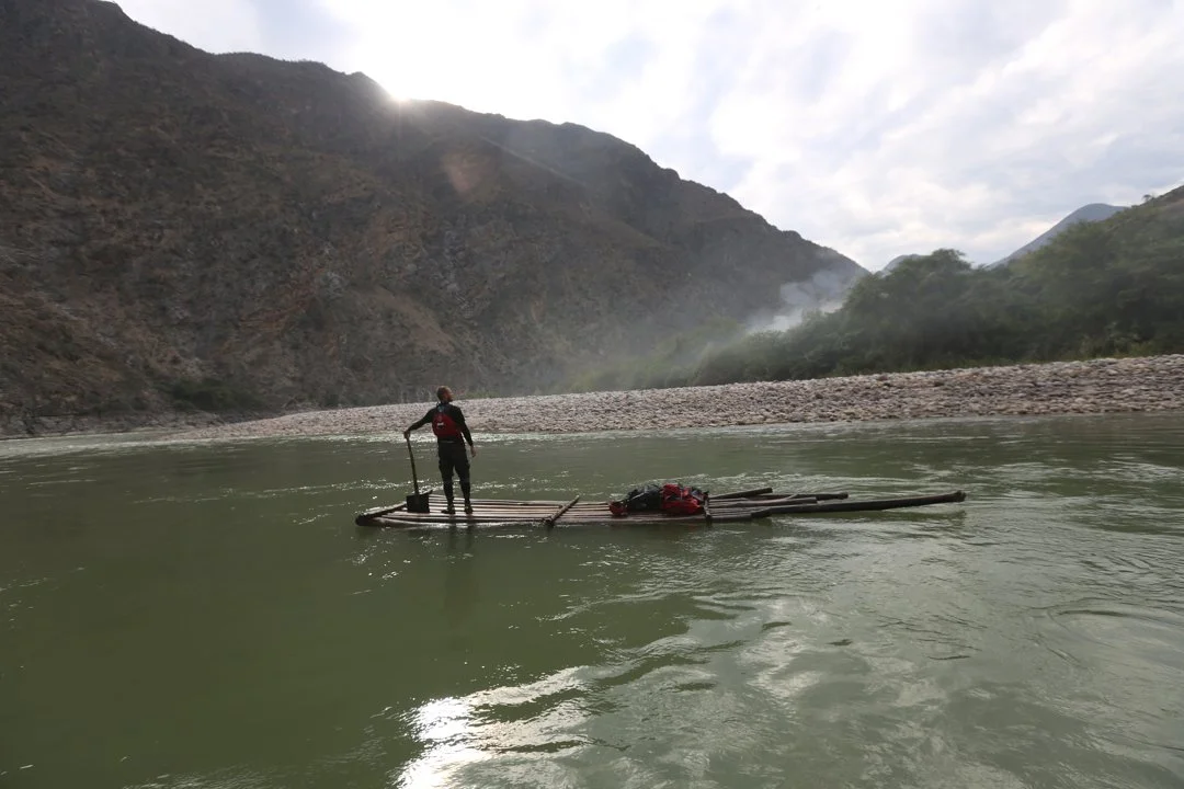 Person standing on makeshift raft with supplies, floating on a river in a mountainous landscape.
