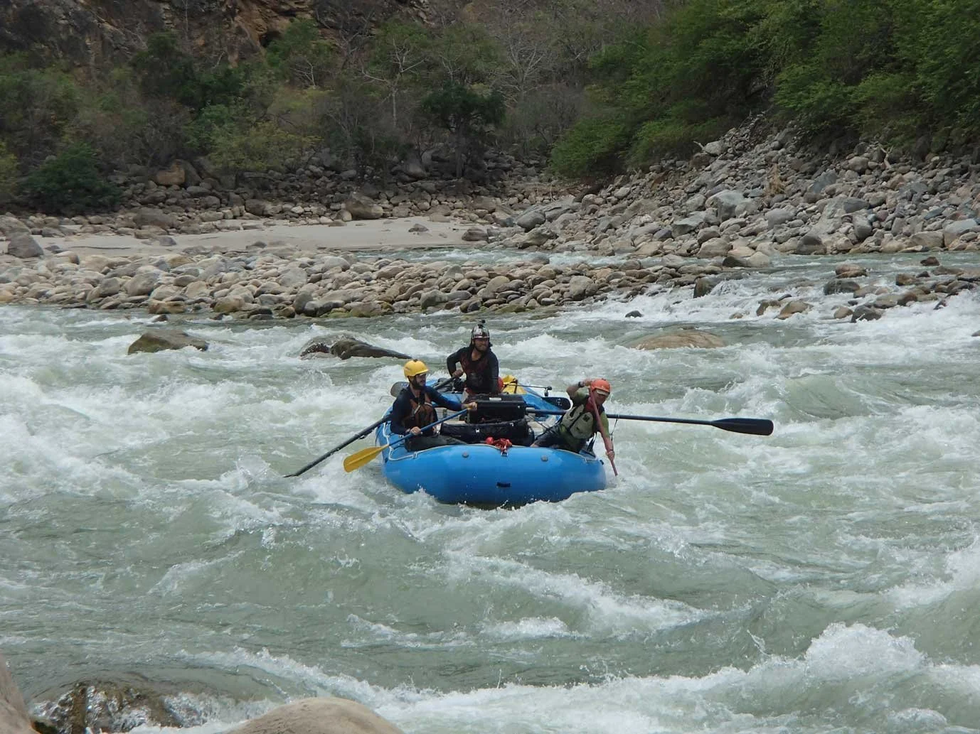 Three people in a blue inflatable raft navigating white water rapids in a river surrounded by rocks and trees.