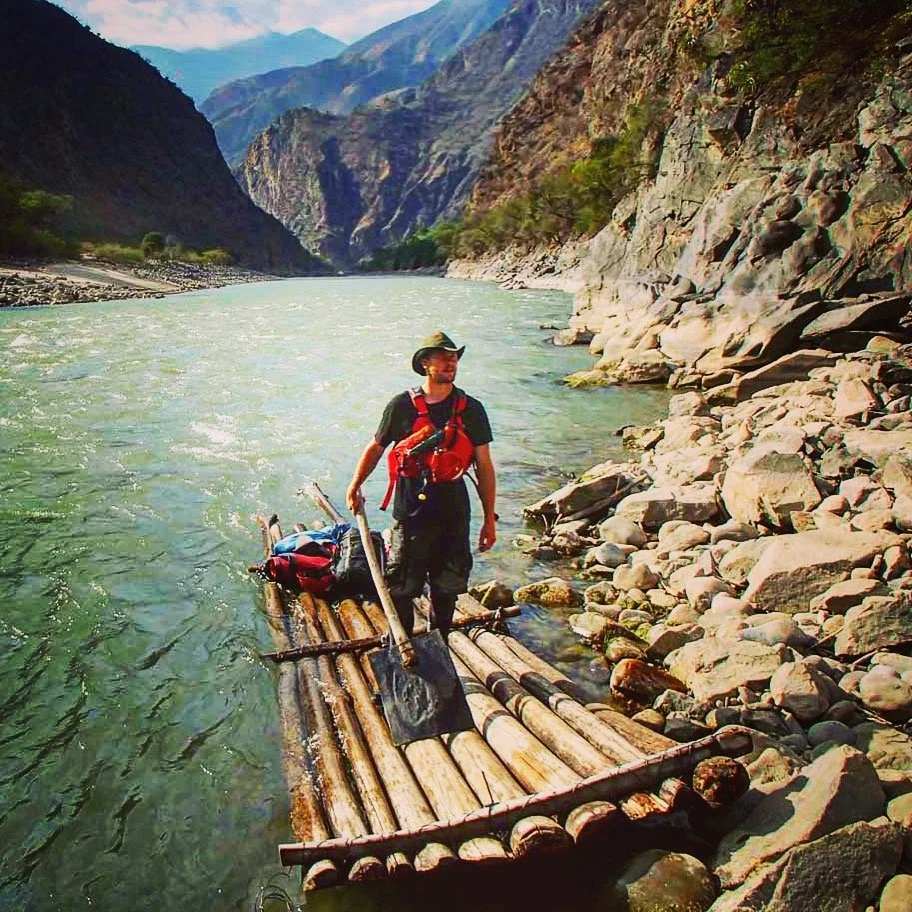A person standing on a bamboo raft in a river surrounded by mountains and rocky shores.