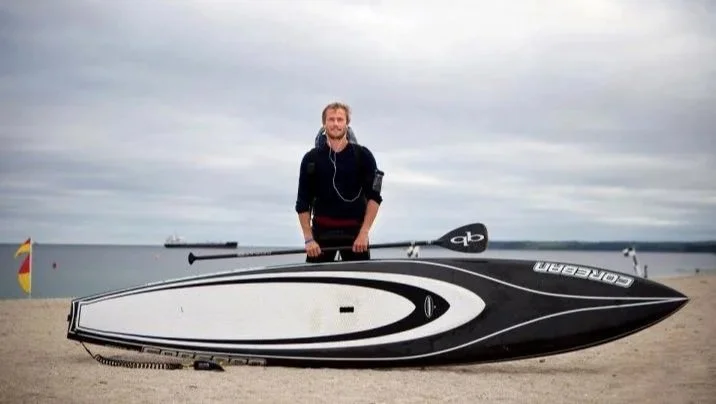 A man standing on a beach next to a large black and white speedboat, with a body of water and ships in the background under a cloudy sky.