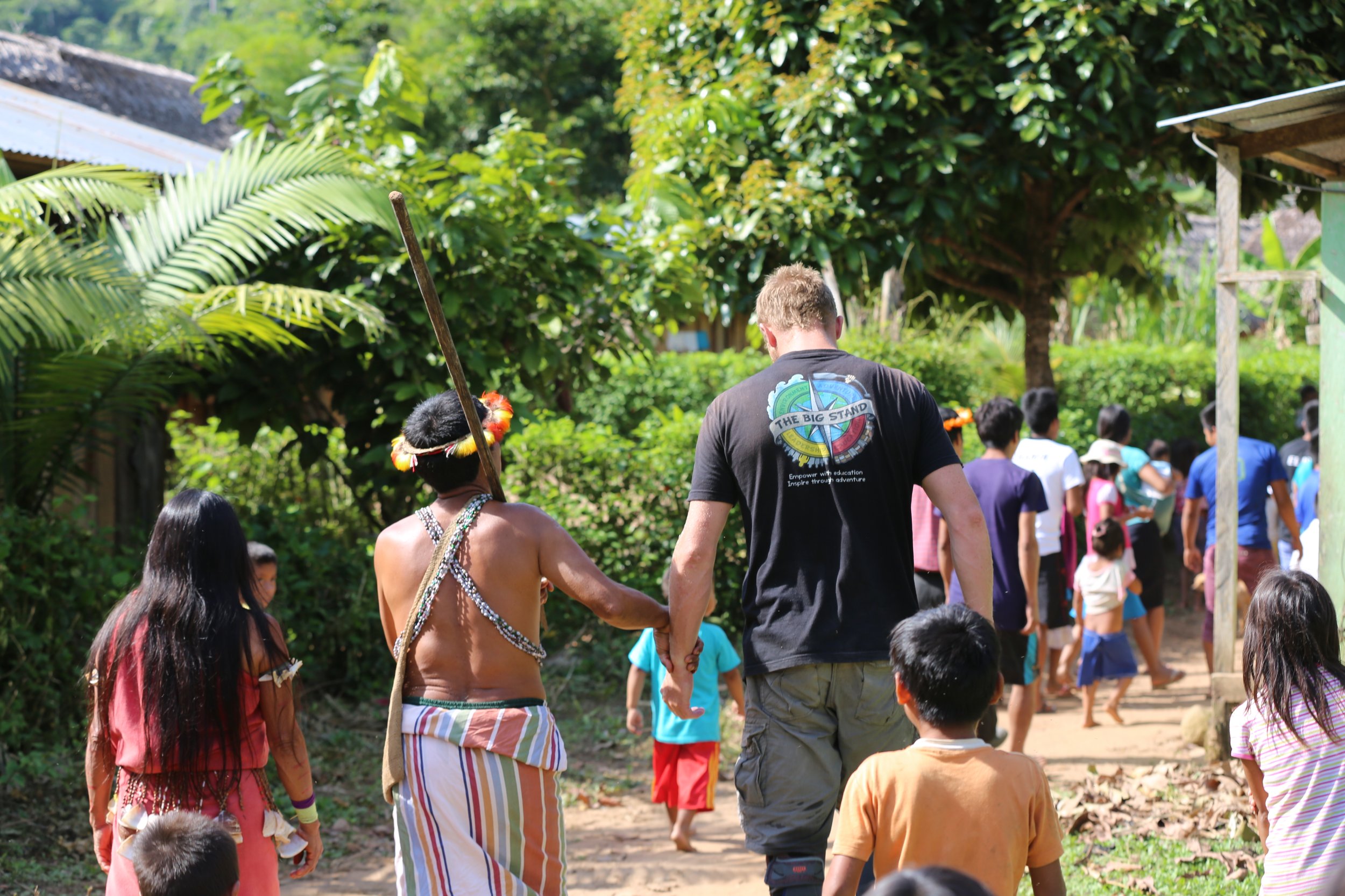 People walking in a lush outdoor setting, including indigenous individuals, children, and young adults, among tropical trees and plants.