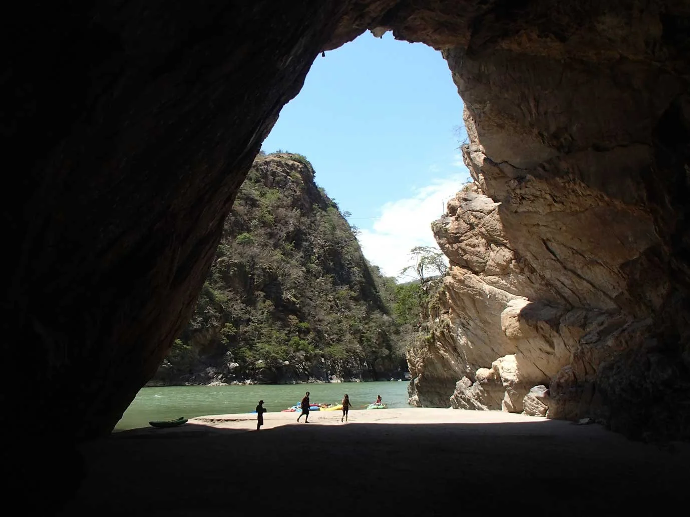 View from inside a large, dark cave looking out at a river with kayakers and people on the sandy shore, surrounded by rocky cliffs and trees under a blue sky.