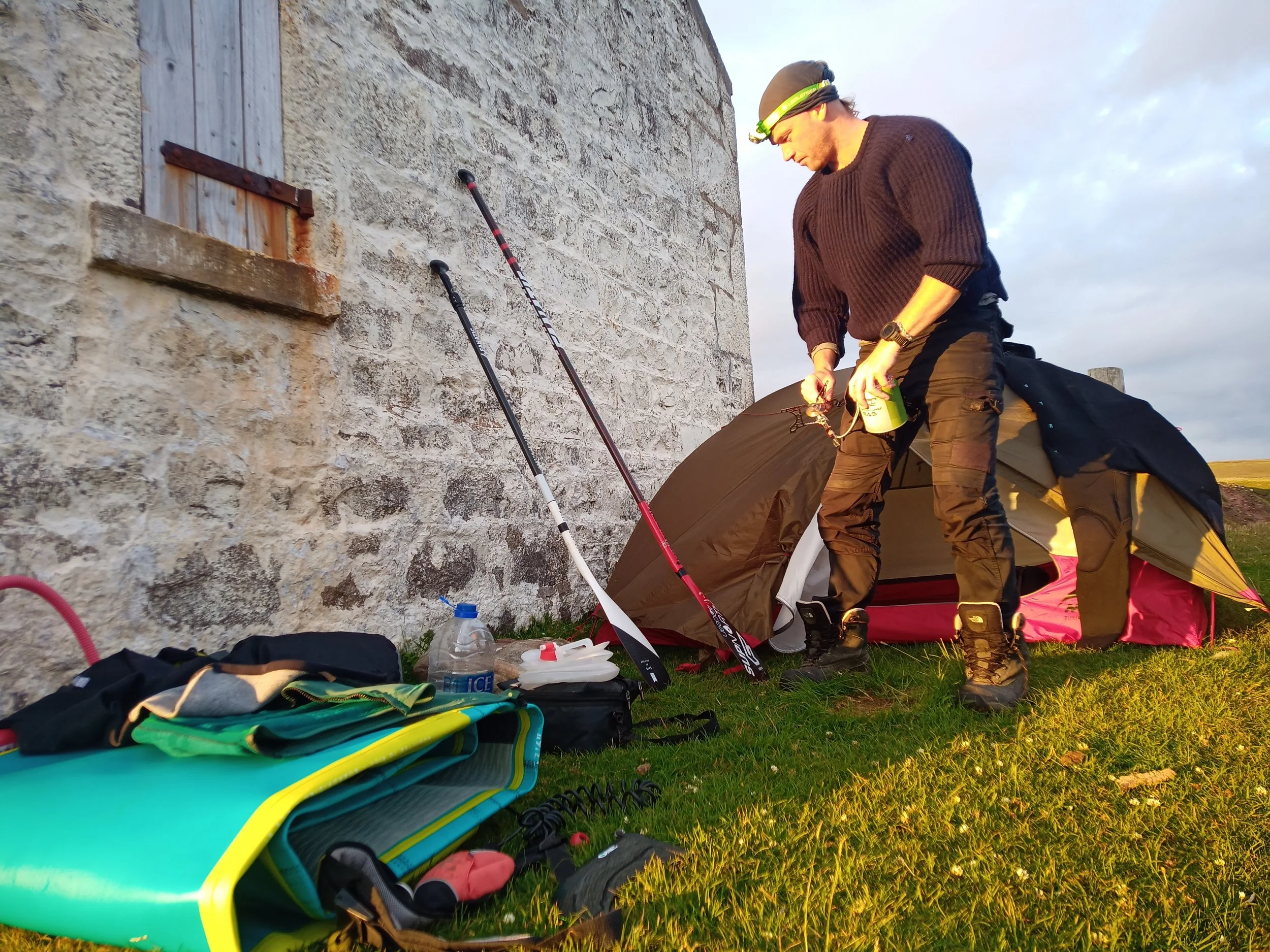 A man in outdoor gear, including a headlamp and brown sweater, sets up camping equipment next to a stone wall with a window shutter. Camping supplies, including a yellow kayak, kayaking paddles, and miscellaneous gear, are spread on the grass nearby.