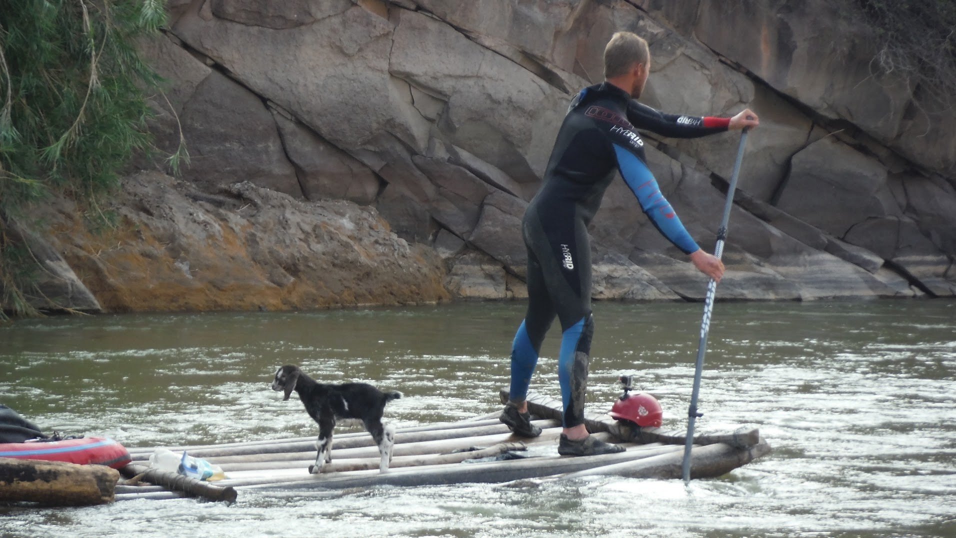 A man wearing a wetsuit standing on a raft in a river, holding a paddle, with a dog standing on the raft nearby, and rocky cliffs in the background.