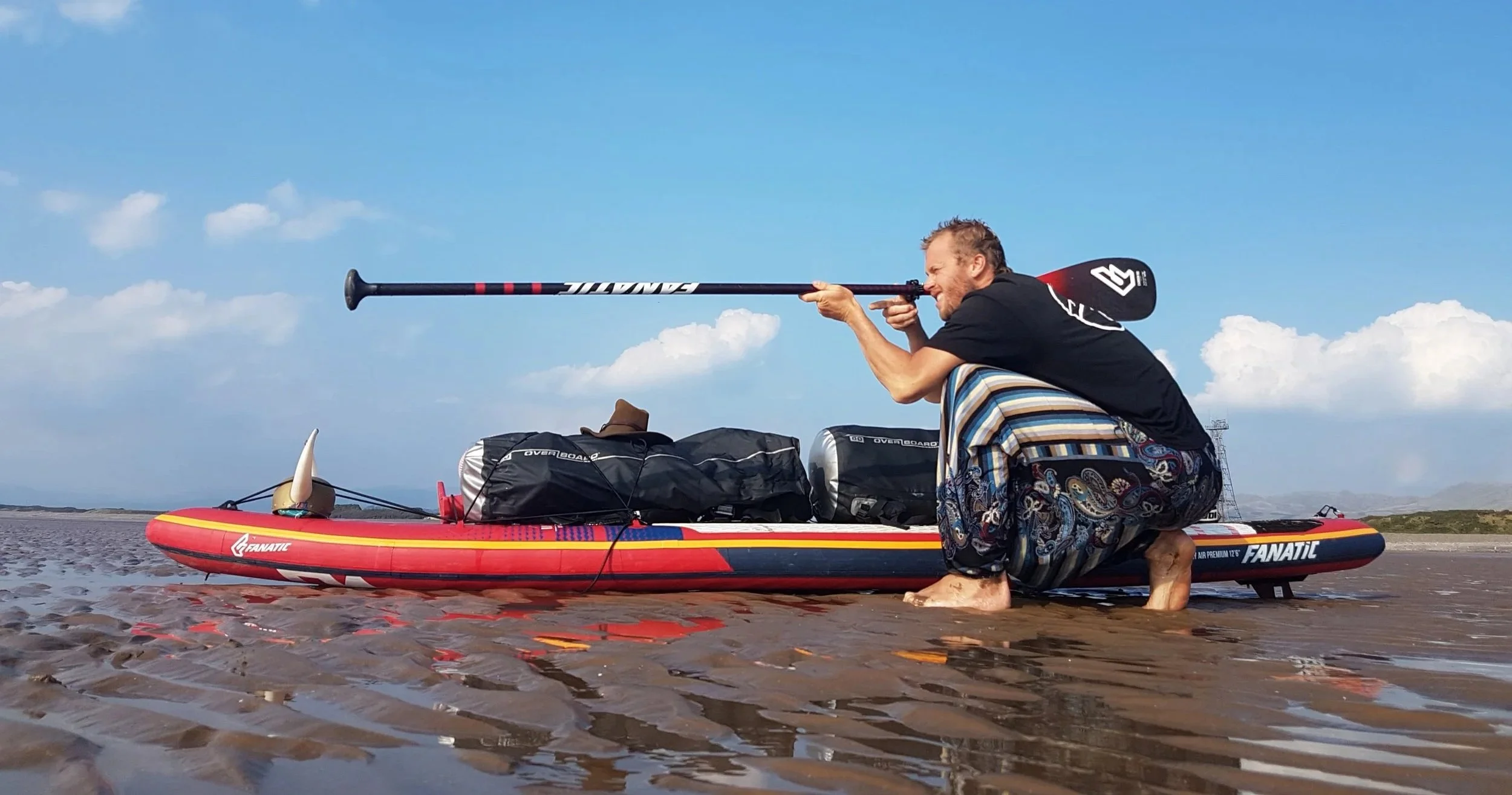 Man kneeling on a paddleboard at the beach, holding a paddle and aiming it like a rifle, with bags and equipment on the paddleboard, ocean and sky with clouds in the background.