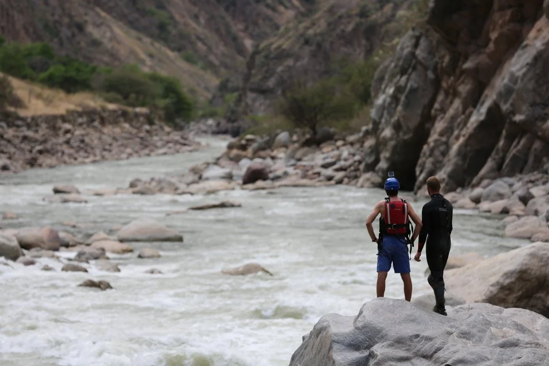 Two men standing on a large rock by a river in a canyon, with rocky cliffs and sparse vegetation in the background.