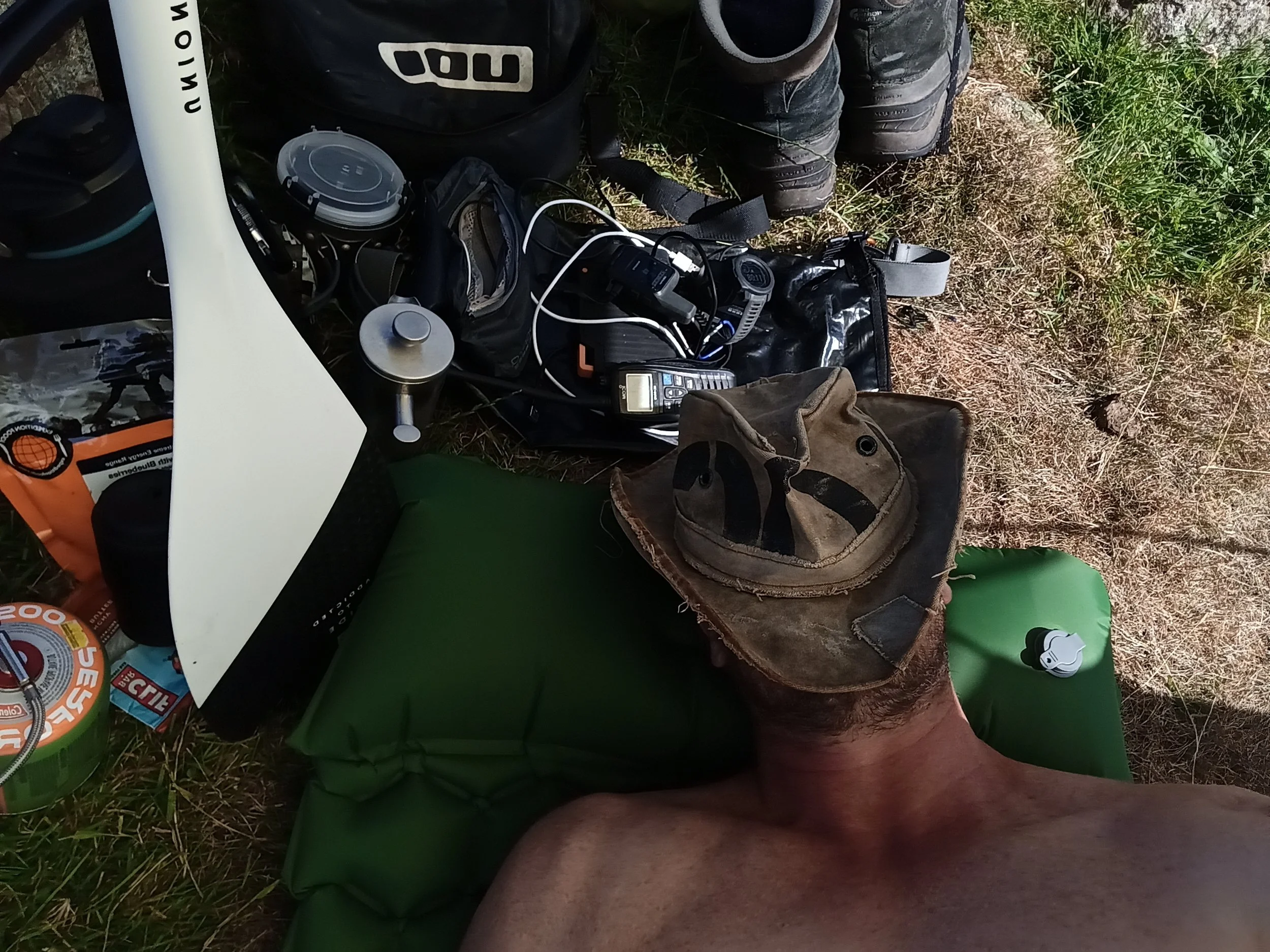 A person lying on the grass with various items surrounding them, including a backpack, a cordless phone, a container, a hat, and some camping or outdoor gear.