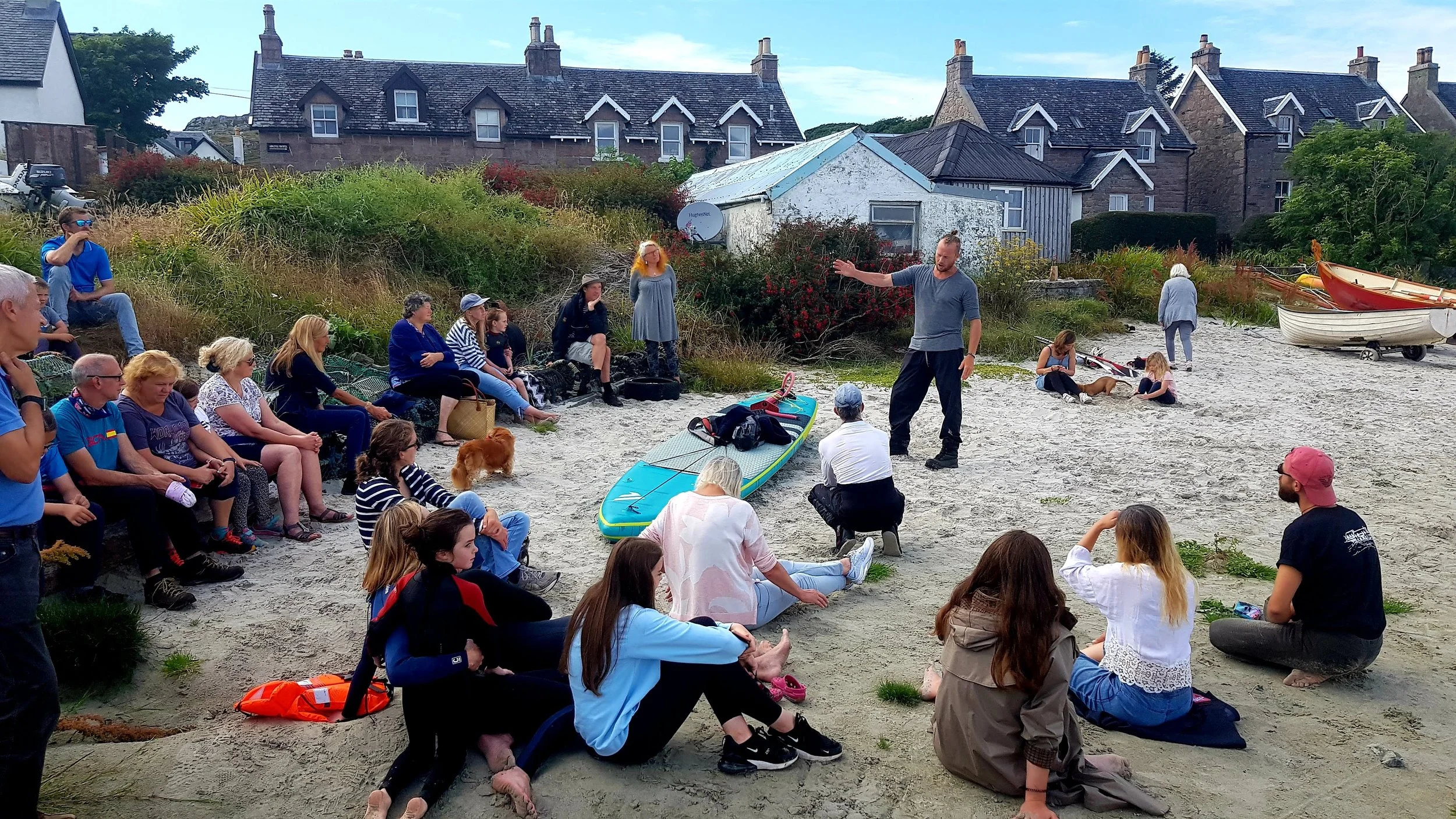 A group of people sitting on the sandy shore listening to a man speaking, with boats on the beach and houses in the background.