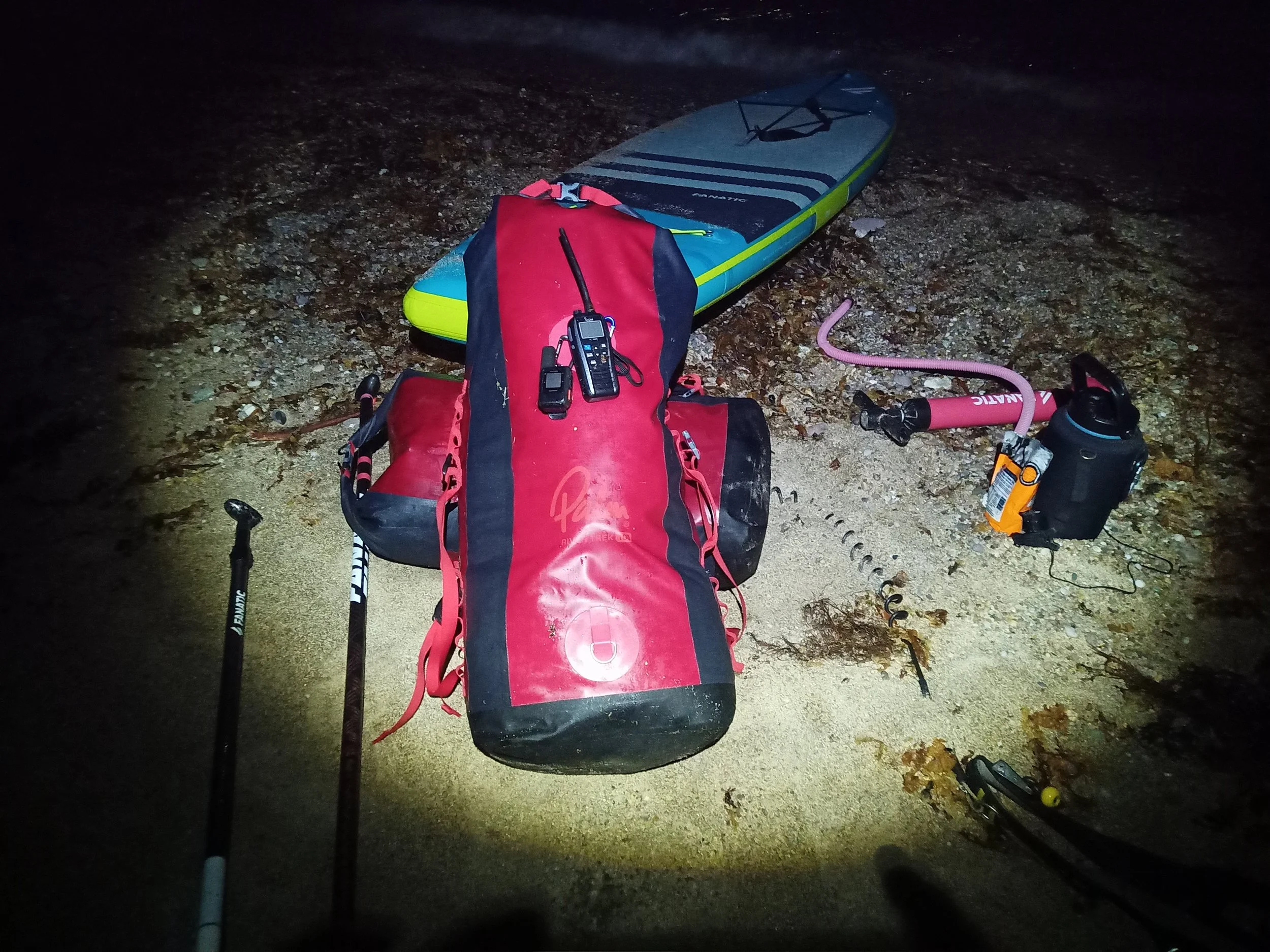 Nighttime scene showing a stand-up paddleboard on rocky ground with a backpack, wireless microphone, and paddle in the foreground.