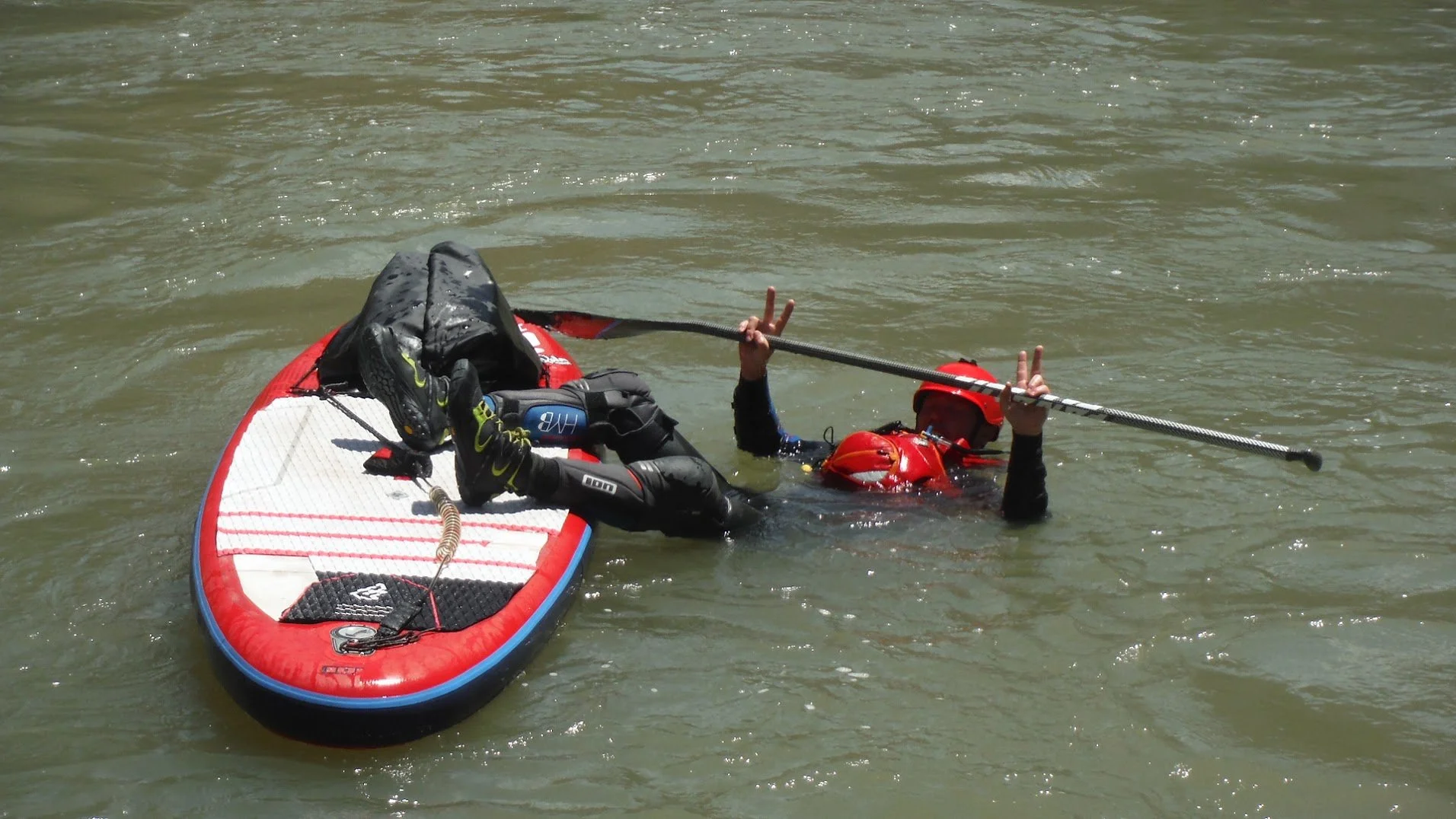 Person in water holding a paddle, lying on a stand-up paddleboard with gear, wearing red helmet and life vest, signaling peace signs with both hands.