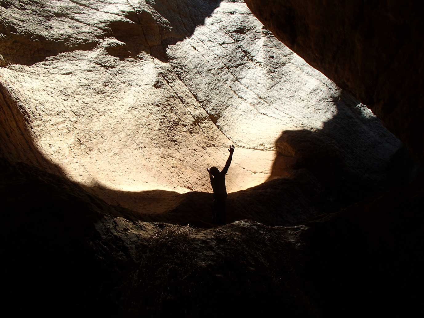 Person standing inside a large, dark cave with one arm raised, lit by sunlight streaming in from above through an opening in the cave ceiling.