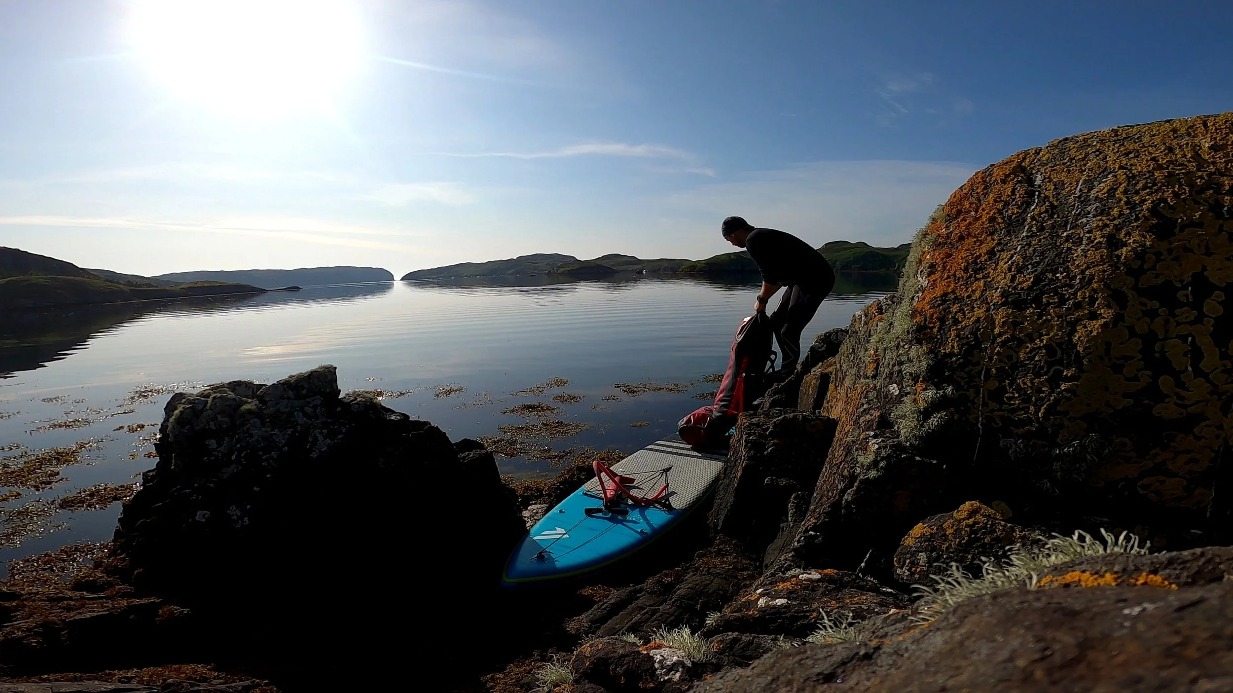 Person preparing to travel with a paddleboard on rocky shoreline with calm water and islands in the distance under sunny sky.