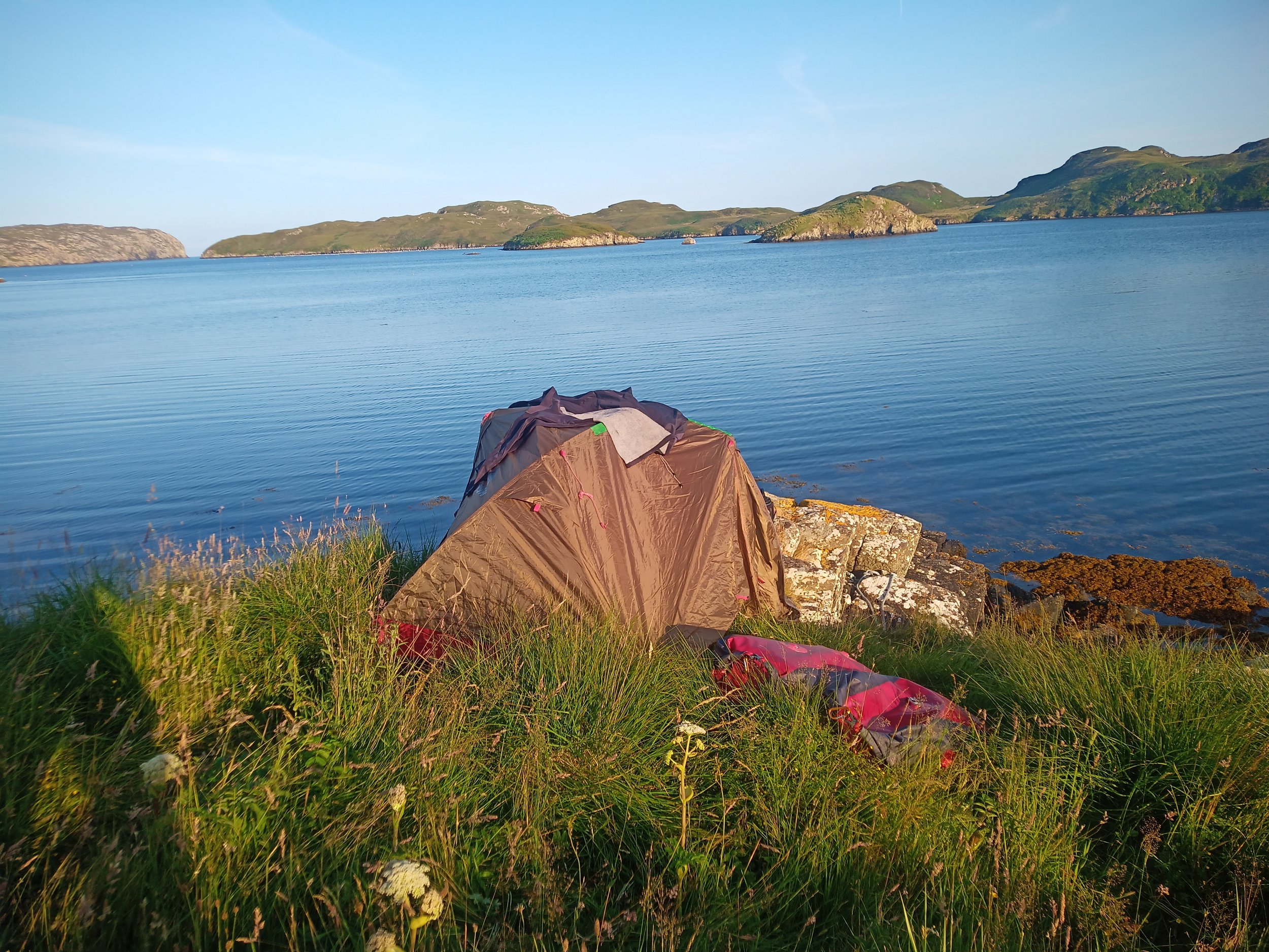 Tent on grassy edge beside a calm body of water with small islands and hills in the background under a clear blue sky.