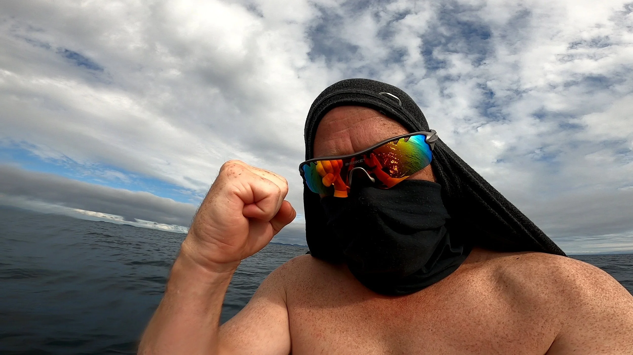 Man on a boat making a fist with his right hand, wearing sunglasses, a black face covering, and a black hat, with water and cloudy sky in the background.