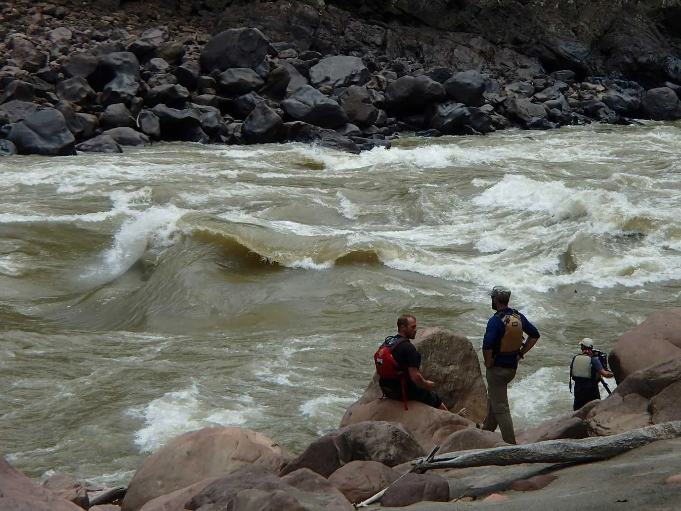 Three people with backpacks exploring a rocky riverbank near turbulent water and rapids.