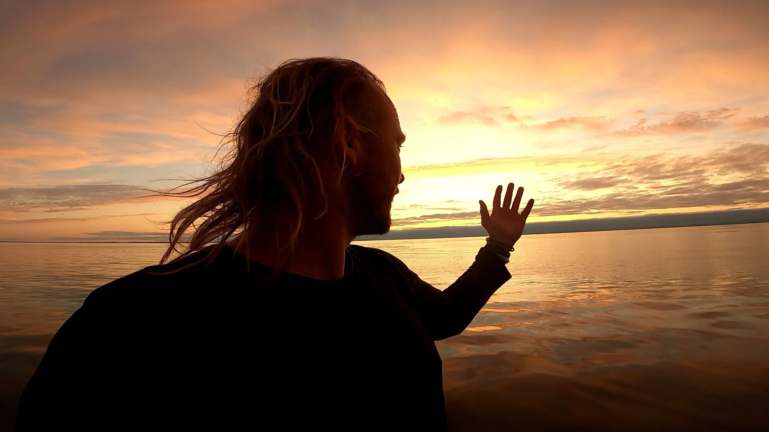 A person with long, wavy hair and a beard is sitting by the water during sunset, with one hand raised towards the sky and the horizon and sky filled with warm, pastel colors.