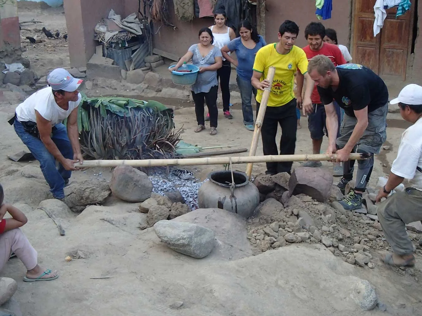 A group of people are gathered outdoors around a large pot over an open fire, with two men using bamboo poles to tend the fire. Others are smiling and watching, with some holding objects, in a rustic setting with mud walls and rocks.