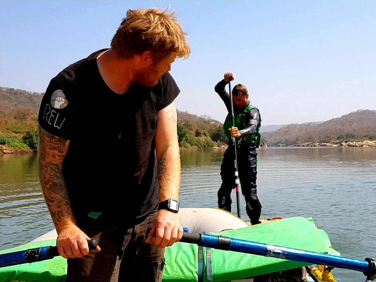Two men on a body of water with hills in the background, one is in the foreground holding a paddle and the other is standing on a paddleboard with a paddle raised.