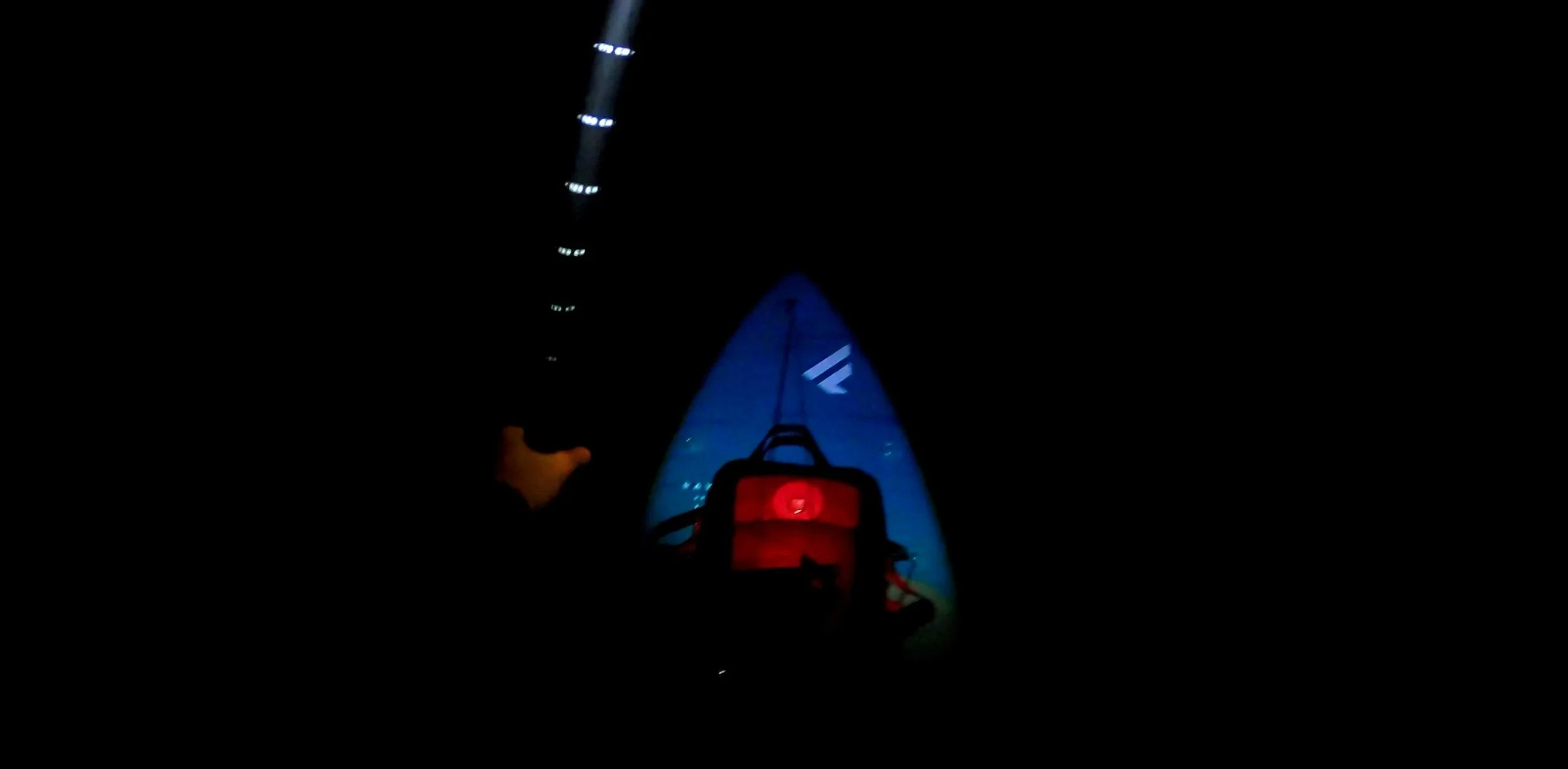 Nighttime photo taken from a first-person perspective of a kayak or small boat, with the boat's bow illuminated in blue and red lights, navigating through dark waters under a sky with a faint starry appearance.