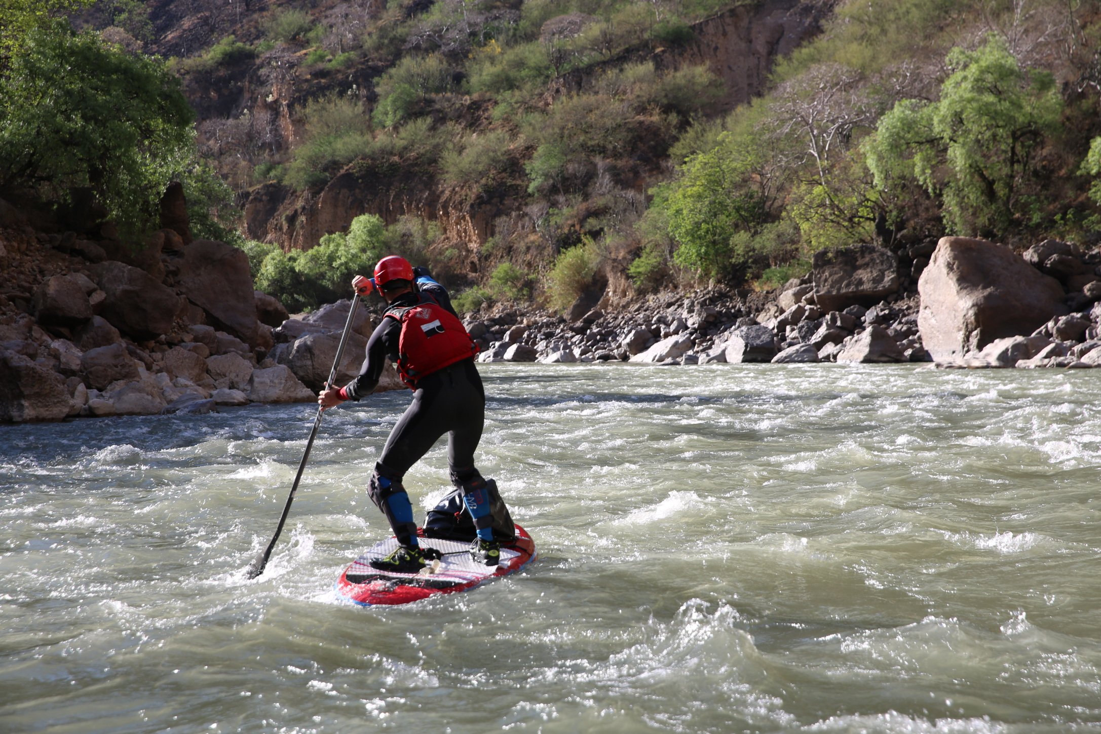 A person in paddleboarding gear, including a red helmet and life vest, is standing on a paddleboard in a river surrounded by rocky banks and lush green trees.