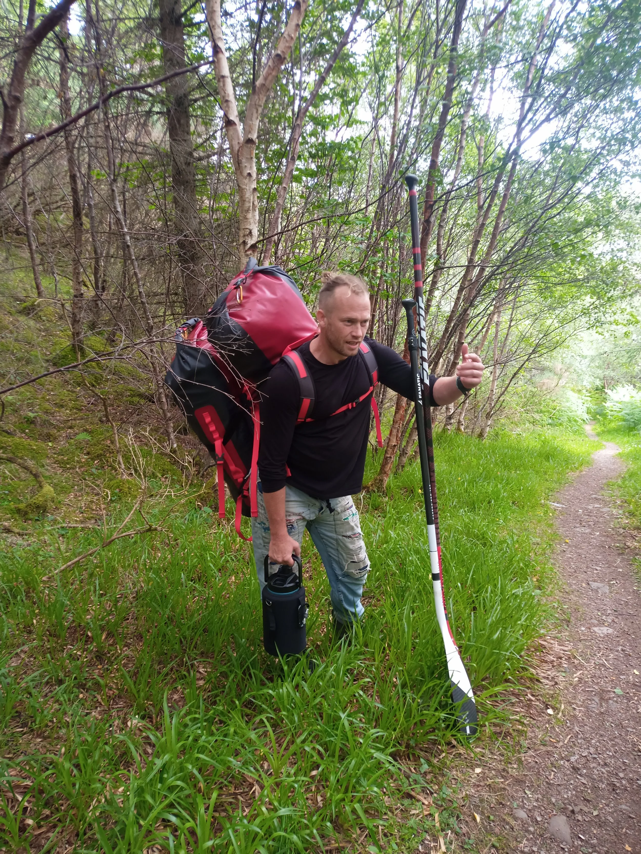 A man carrying a large backpack and hiking poles on a trail through a wooded area.