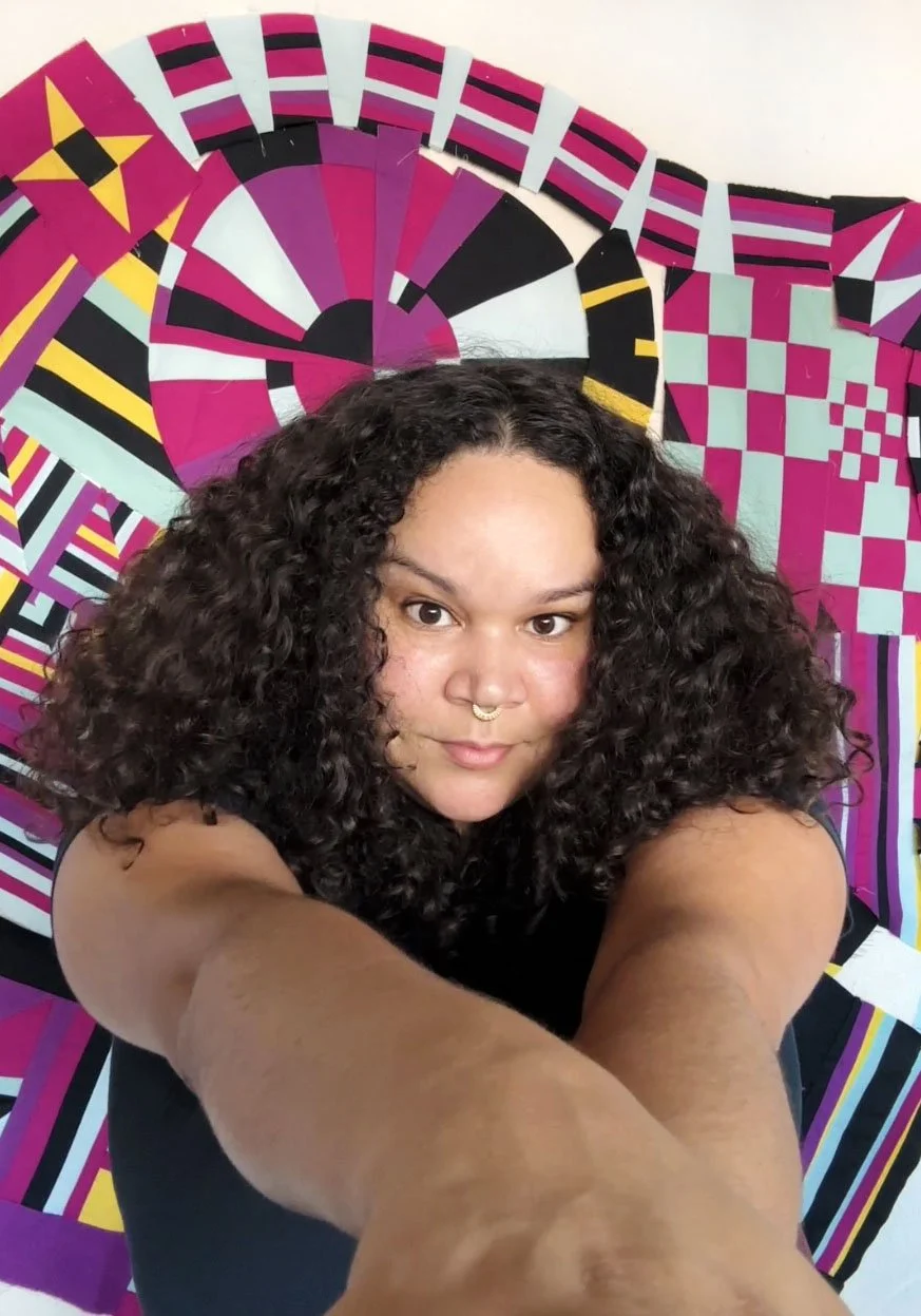 A woman with curly dark hair and a septum piercing taking a selfie in front of a colorful, geometric patterned background.