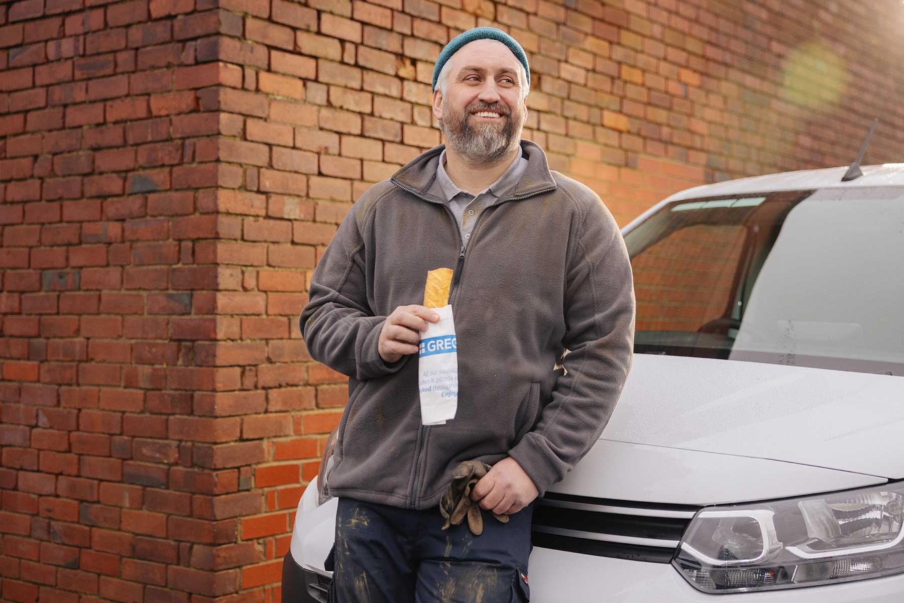  Man with beard and a beanie hat is enjoying a `Greggs Sausage Roll 