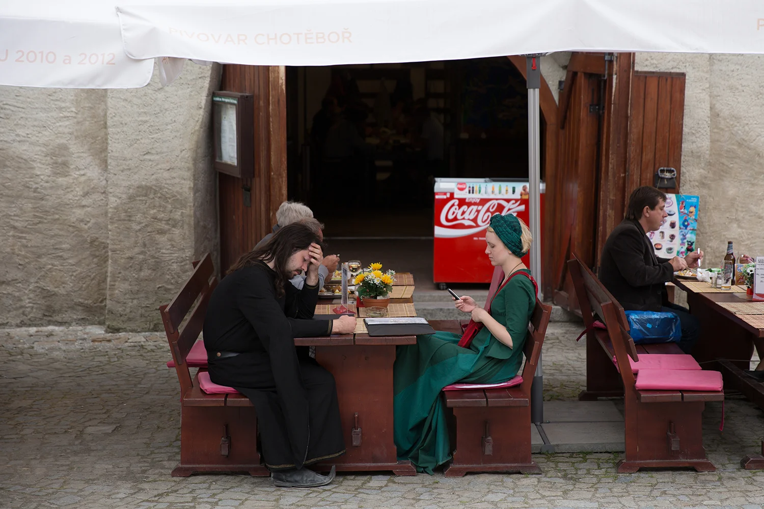 A documentary photograph of two cesky krumlov residents eating lunch and texting while wearing costumes from historical time in Czech Republic