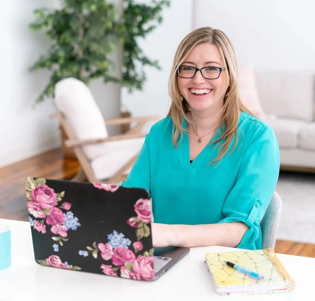 Smiling woman with blonde hair and glasses working on a laptop at a desk.
