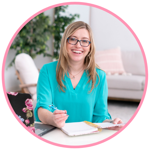 A woman with blonde hair and glasses smiling while sitting at a desk, writing in a notebook with a pen, in a bright, modern living room.
