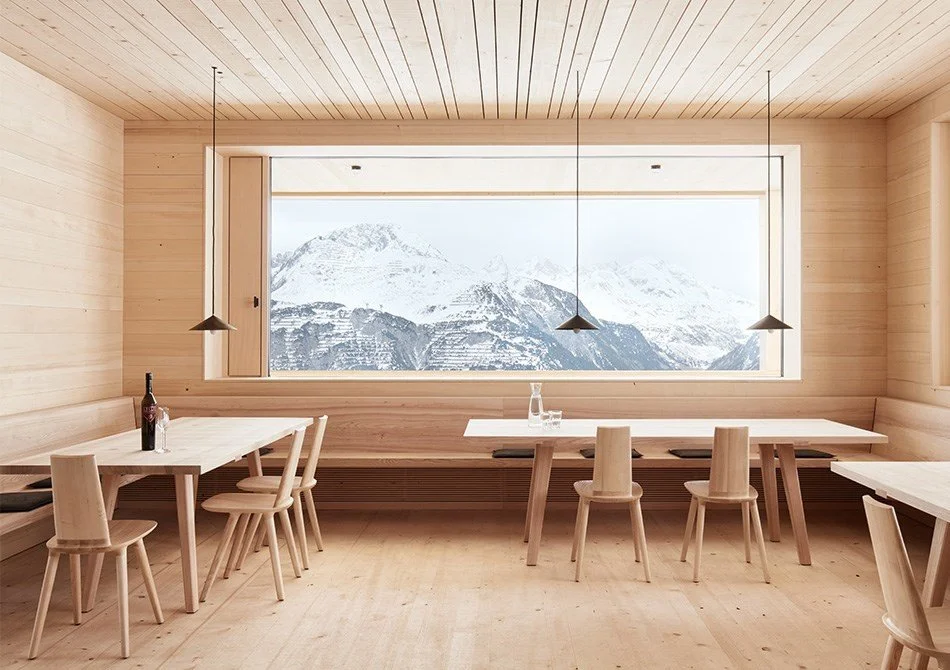 Intérieur d'une salle à manger en bois clair avec vue sur des montagnes enneigées à travers une grande fenêtre