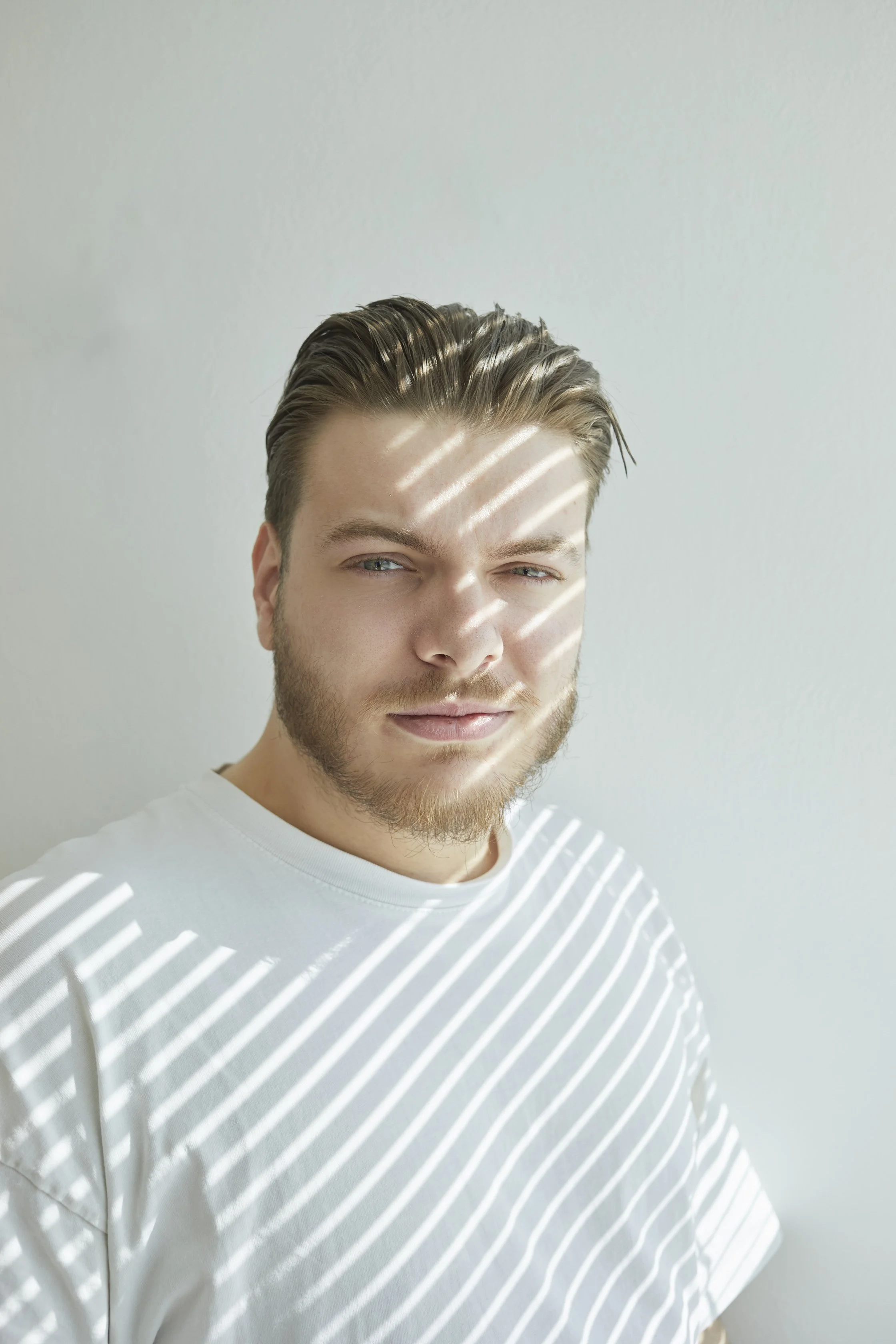 Jeune homme avec cheveux bruns, barbe légère, portant un t-shirt blanc, sous la lumière du soleil avec des rayures de lumière sur son visage et son corps.